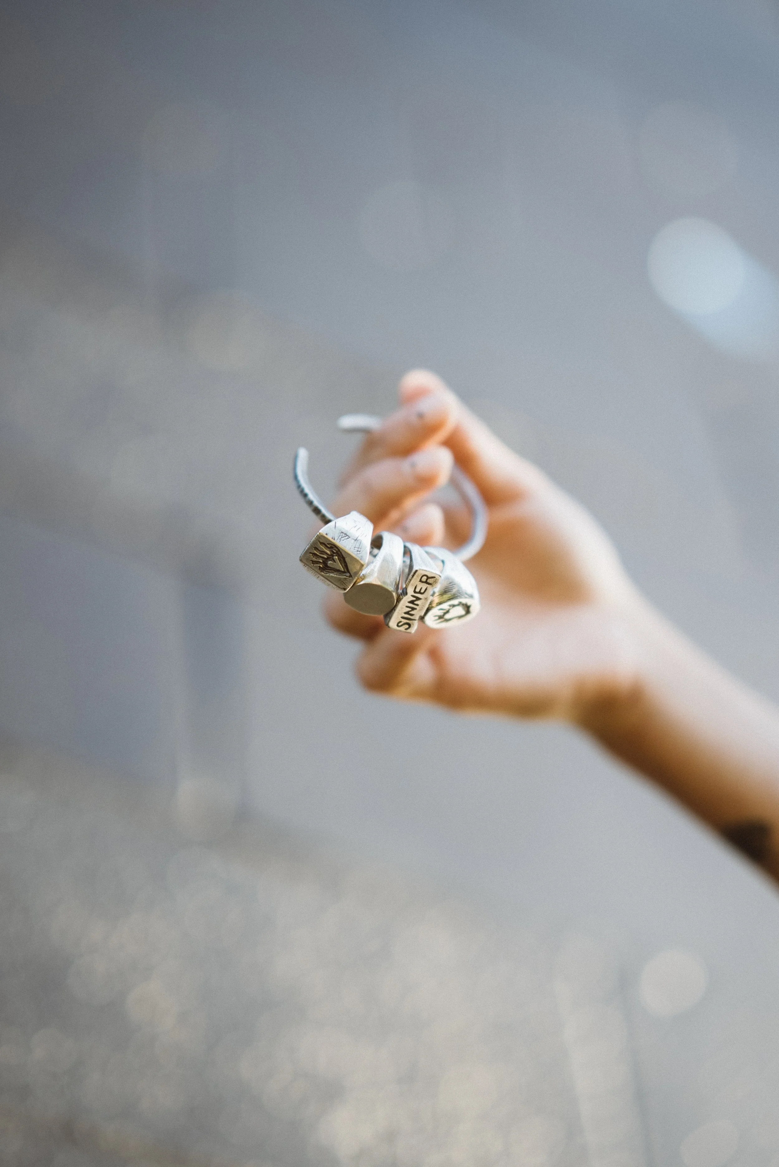 A hand holding silver rings, one with the inscription SINNER, against a blurred background.