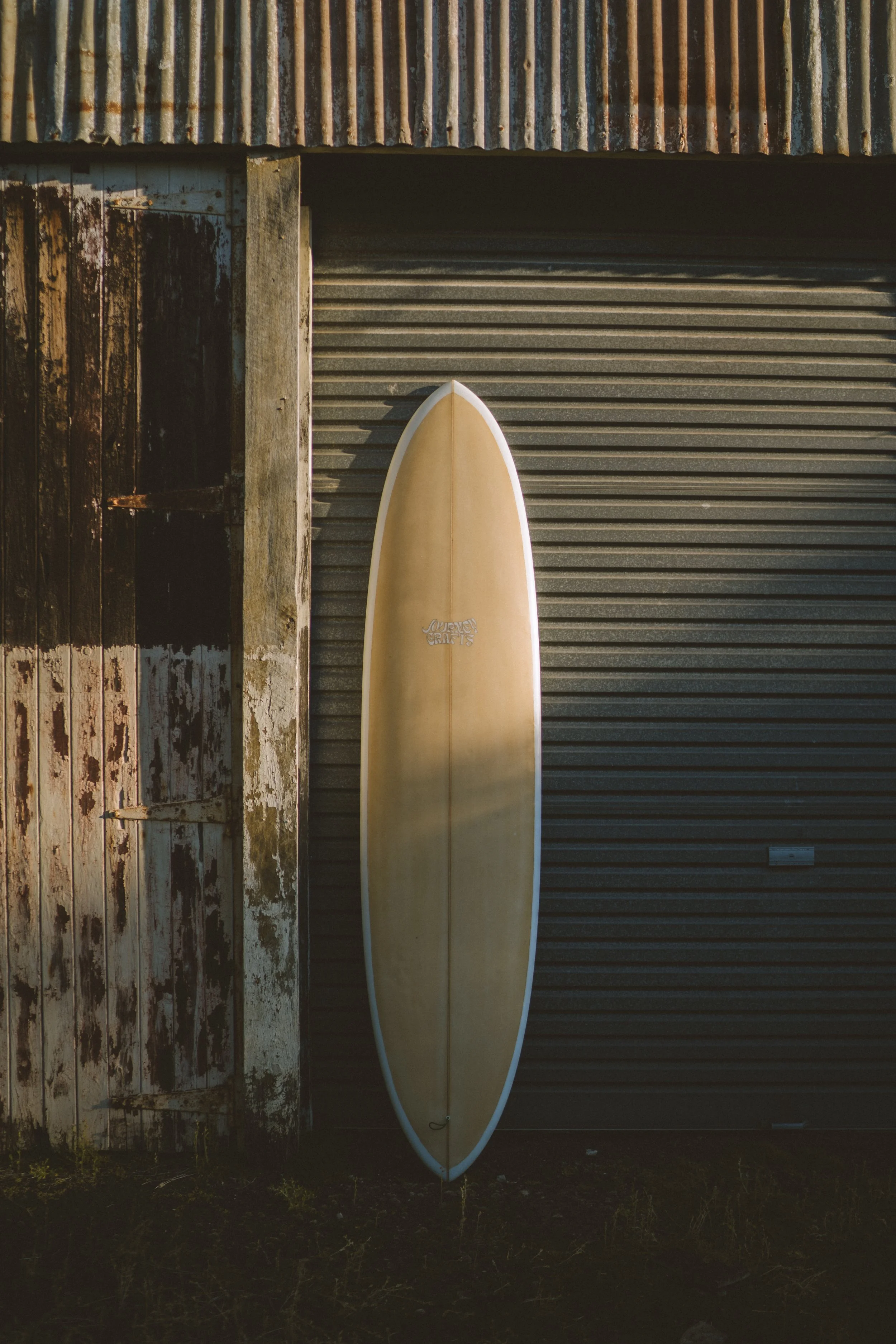 A surfboard leaning against a rusty, weathered metal shed with a corrugated metal door.