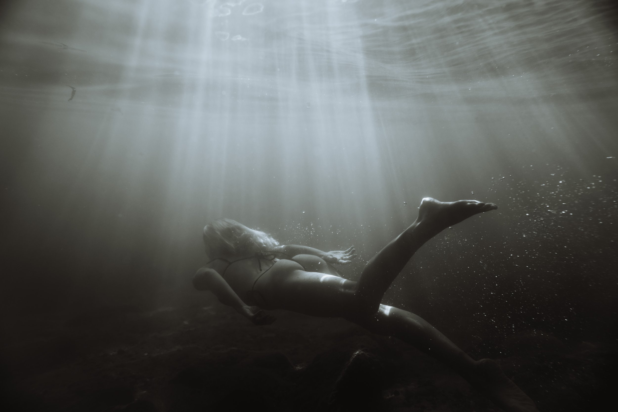 A woman swimming underwater, illuminated by rays of sunlight coming from above, with her head tilted back and her arms extended.