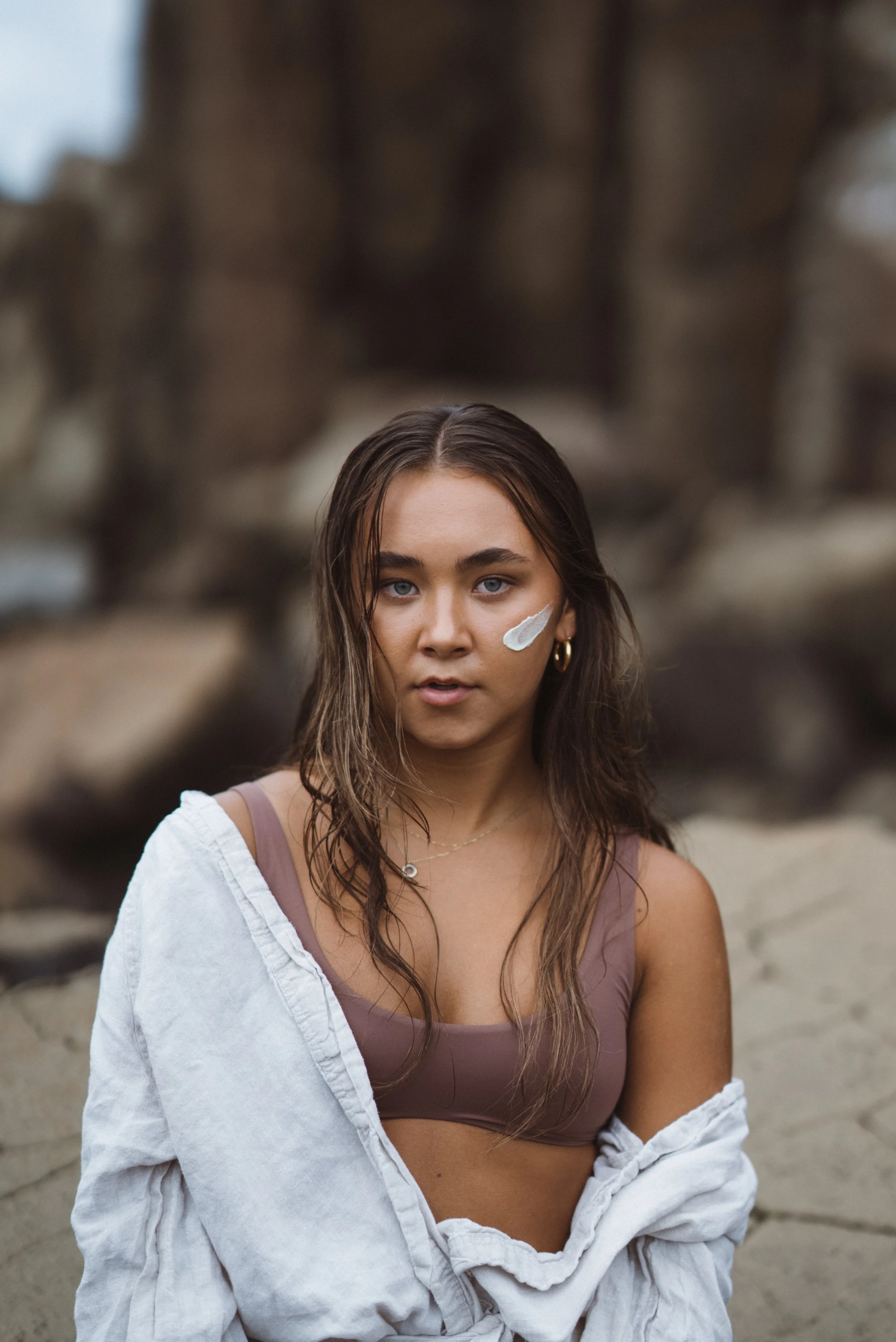Young woman with wet hair and a cream on her face sitting outdoors on a rocky surface, wearing a mauve sports bra and an open white shirt.