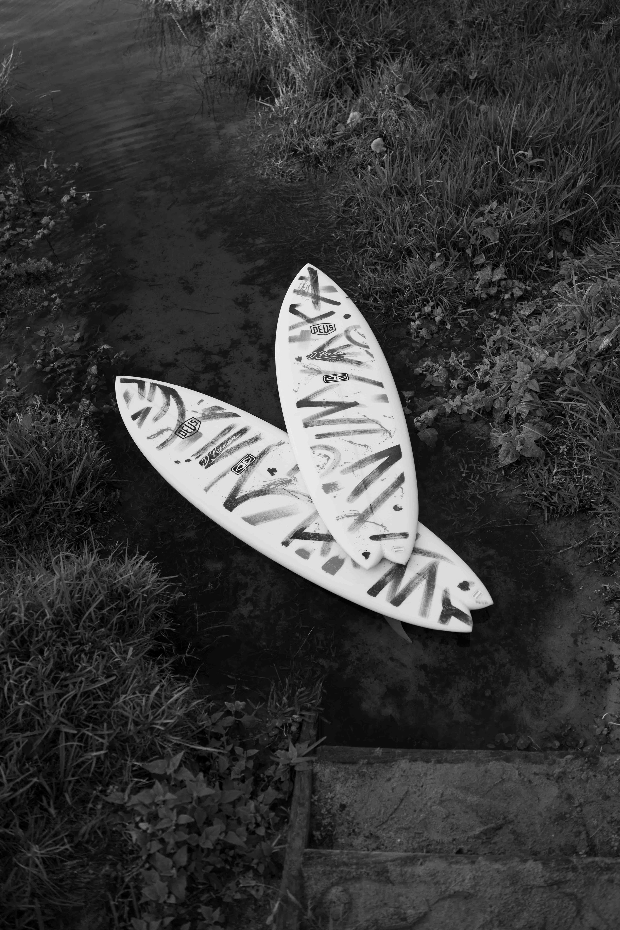 Two used white skateboards with graffiti-style designs lying on the ground near a water stream with grass and plants around.
