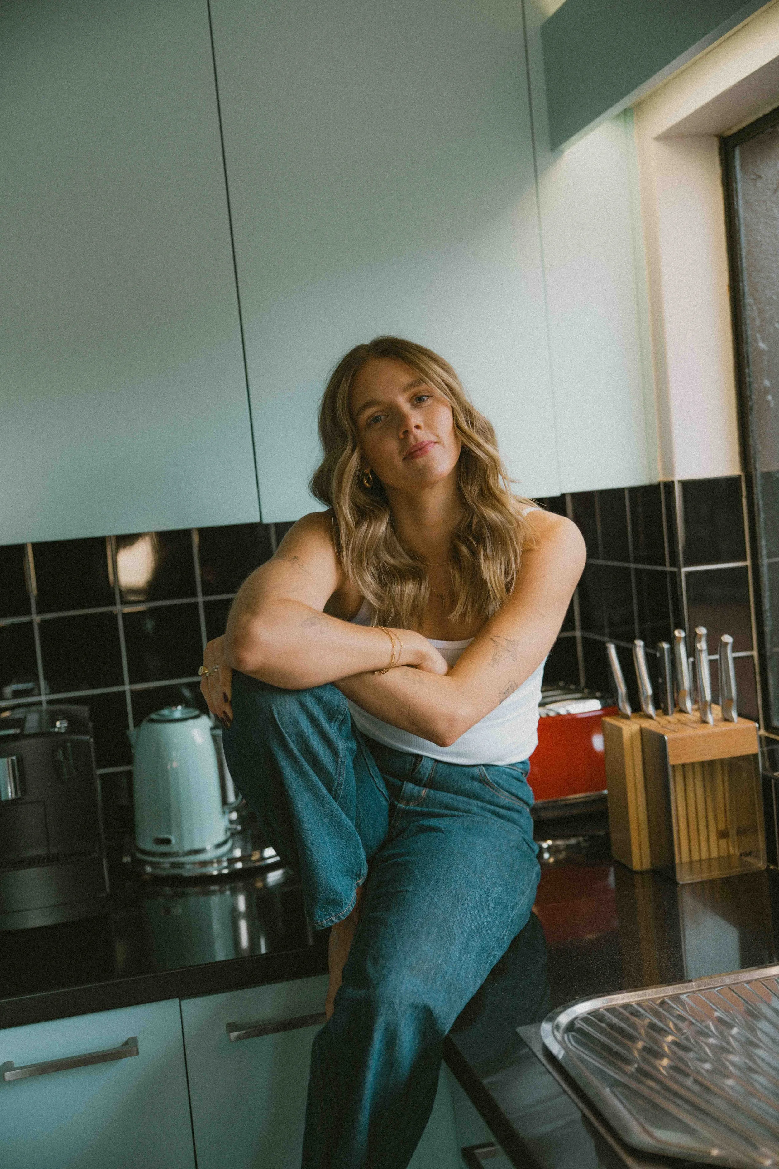 Young woman with wavy hair sitting on a kitchen counter, looking at camera with relaxed expression, white tank top, blue jeans, in modern kitchen with black backsplash, teal cabinets, knife set, toaster, kettle, and window.