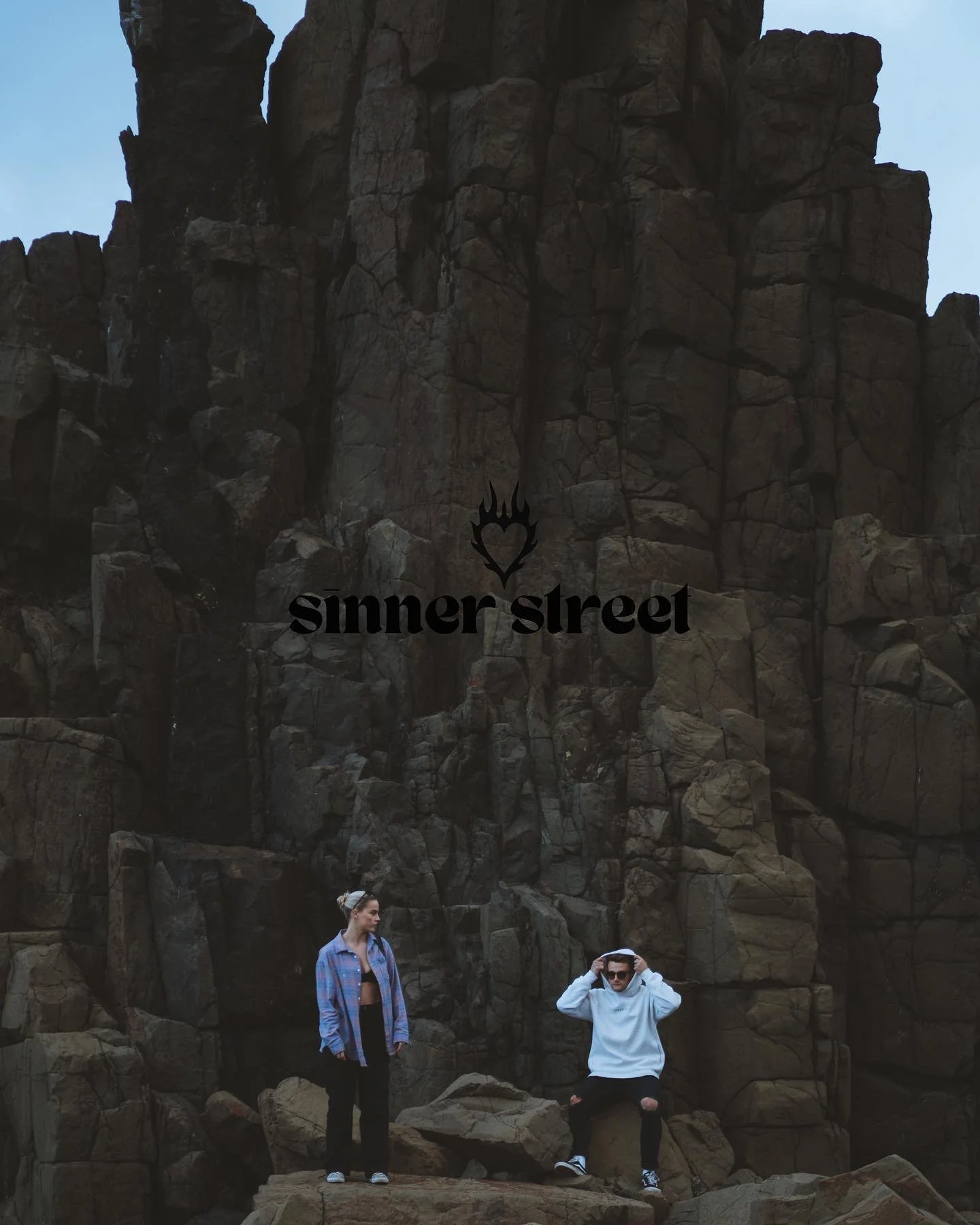 Two people standing and sitting on rocks in front of a large rocky cliff in a natural outdoor setting.