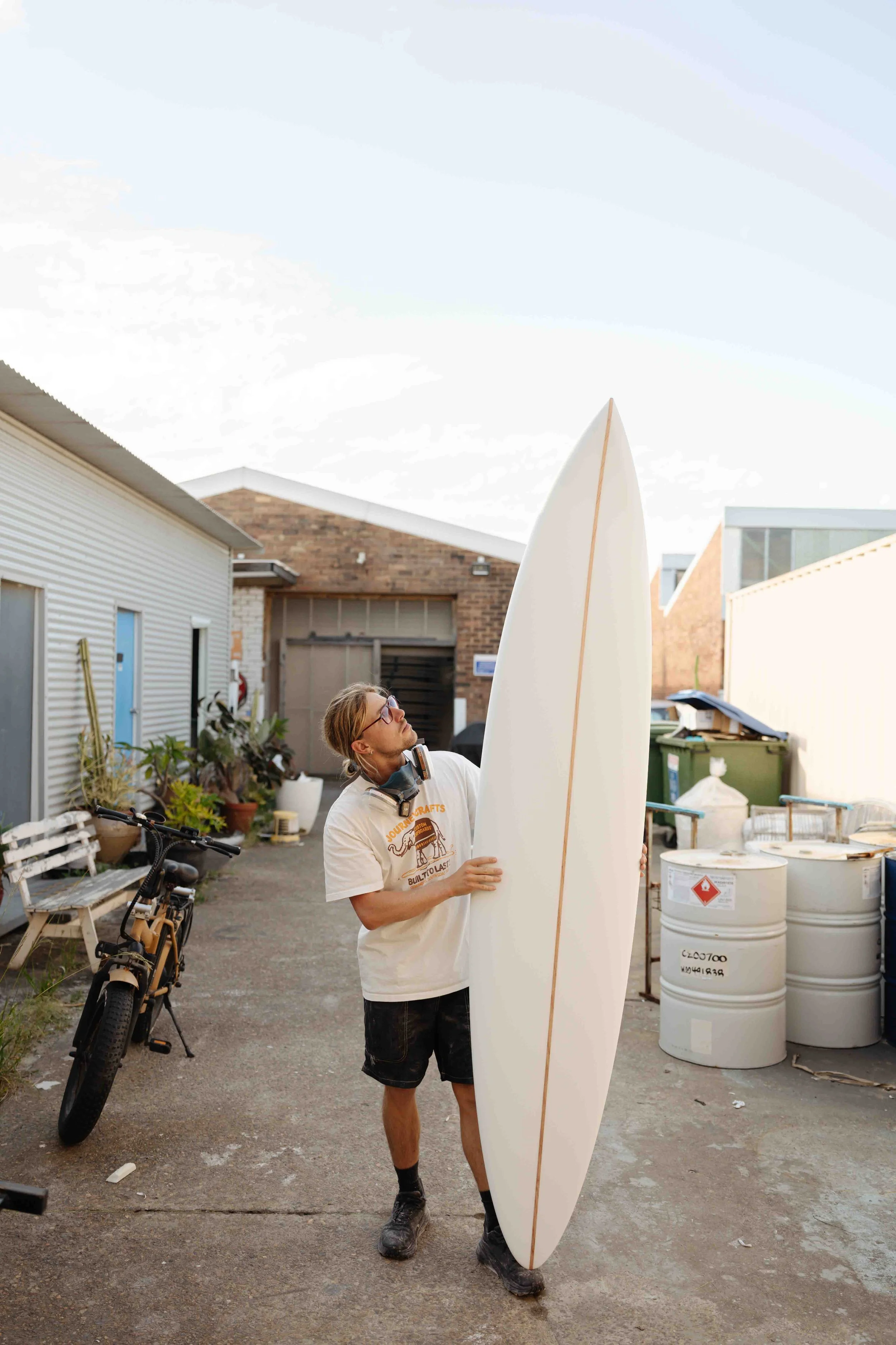 A man standing outdoors, holding a surfboard upright, looking up at it. He is wearing glasses, a white T-shirt, shorts, and has headphones around his neck. There is a bicycle, potted plants, and barrels in the background.