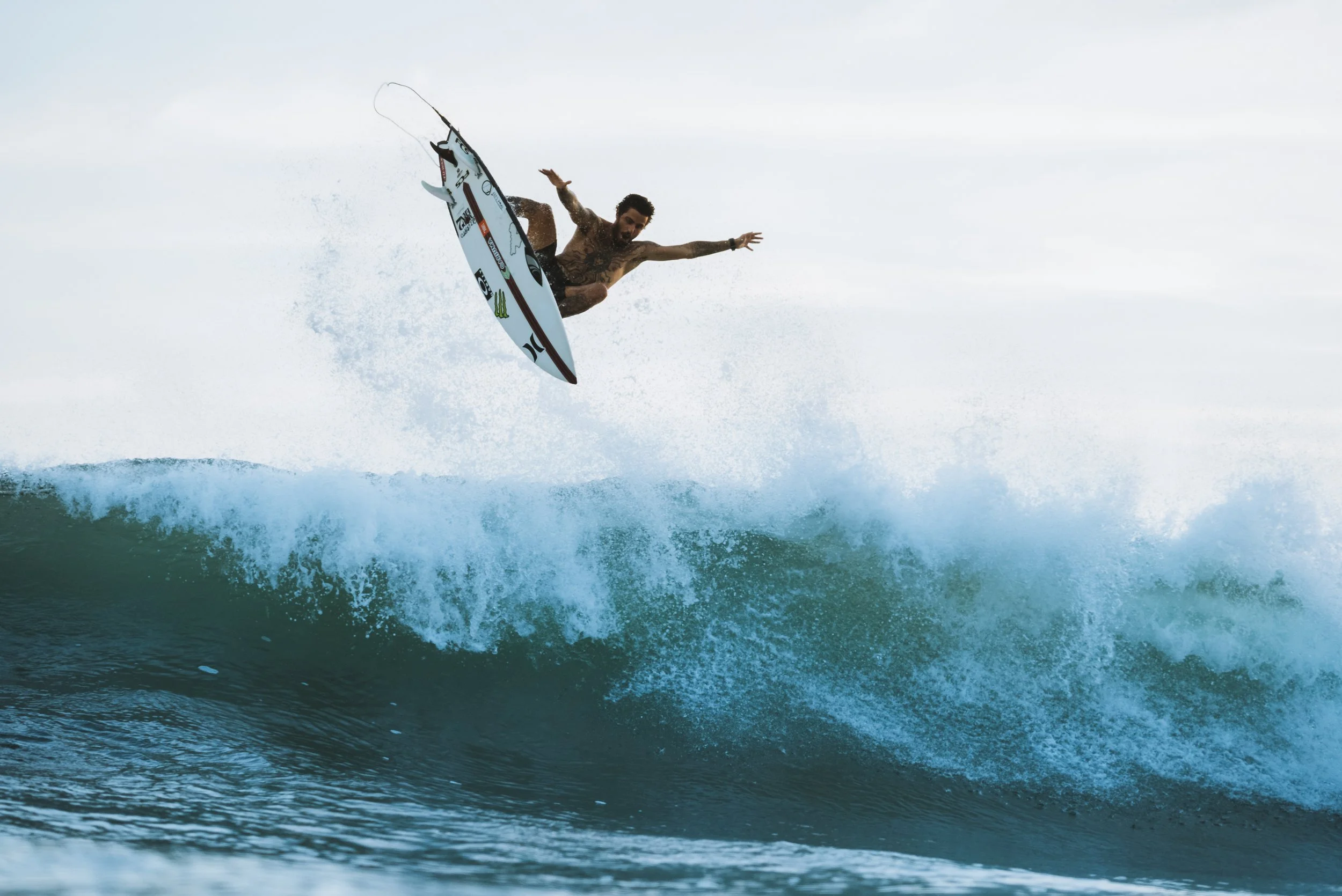 A male surfer is soaring above a wave on his surfboard during the daytime, with arms outstretched and a focused expression.