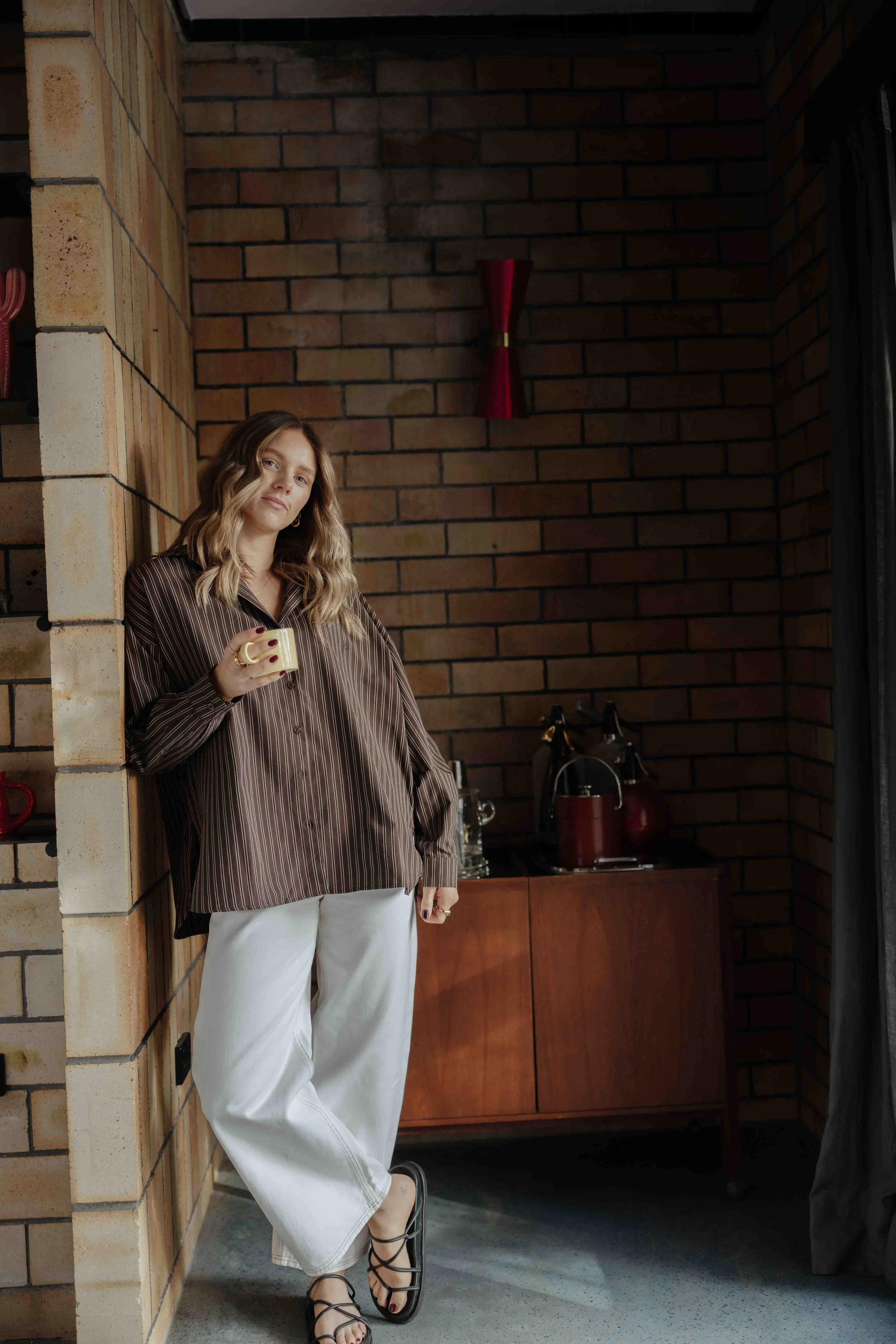 Woman with wavy hair in a brown striped blouse and white pants holding a mug, standing in a modern room with brick walls and a wooden cabinet, with a red vase on the wall behind her.