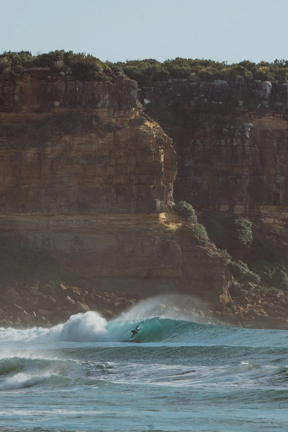 A person surfing on a wave near a rocky coastline with cliffs and sparse vegetation.