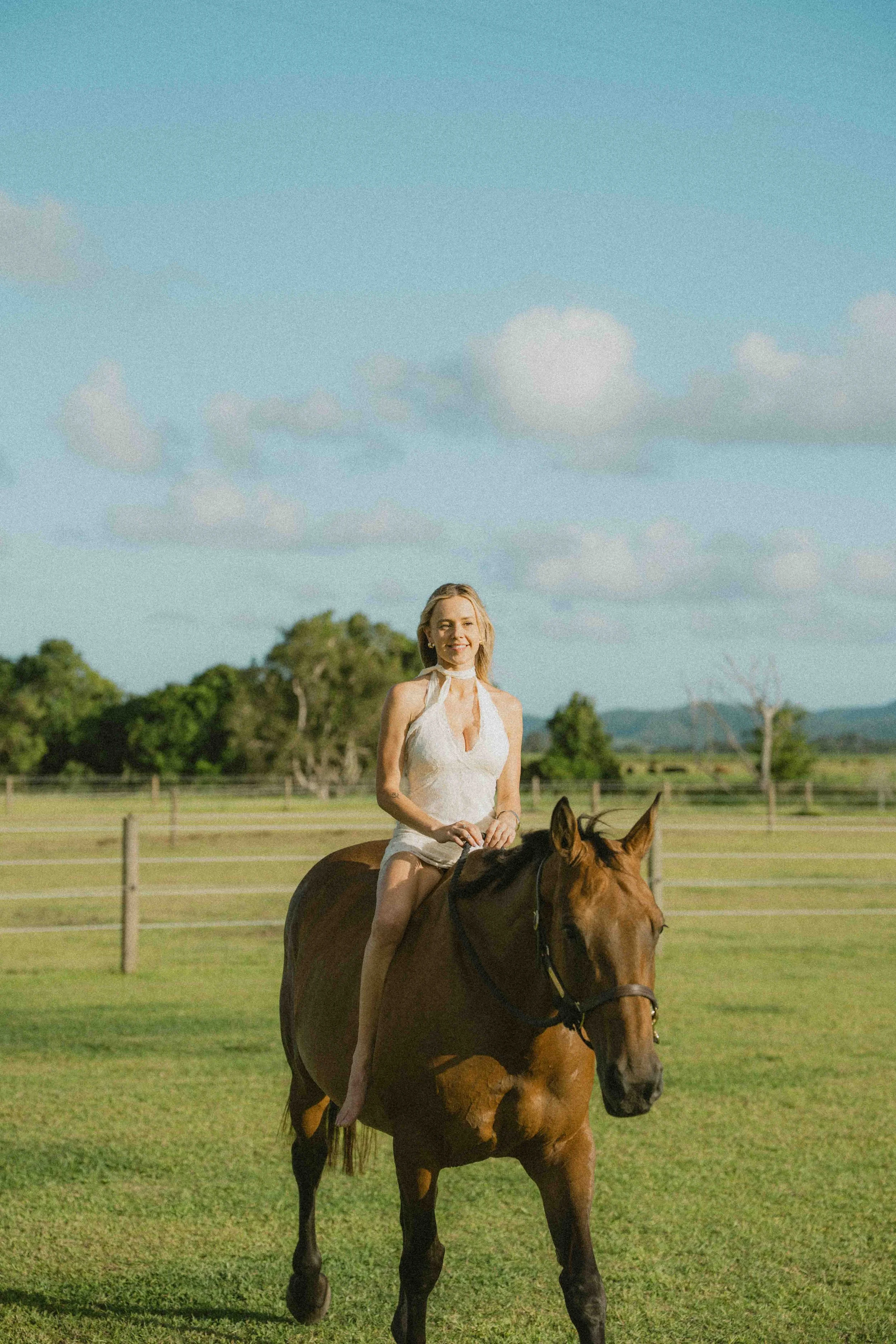 A woman in white dress riding a brown horse in a green field under a partly cloudy sky.