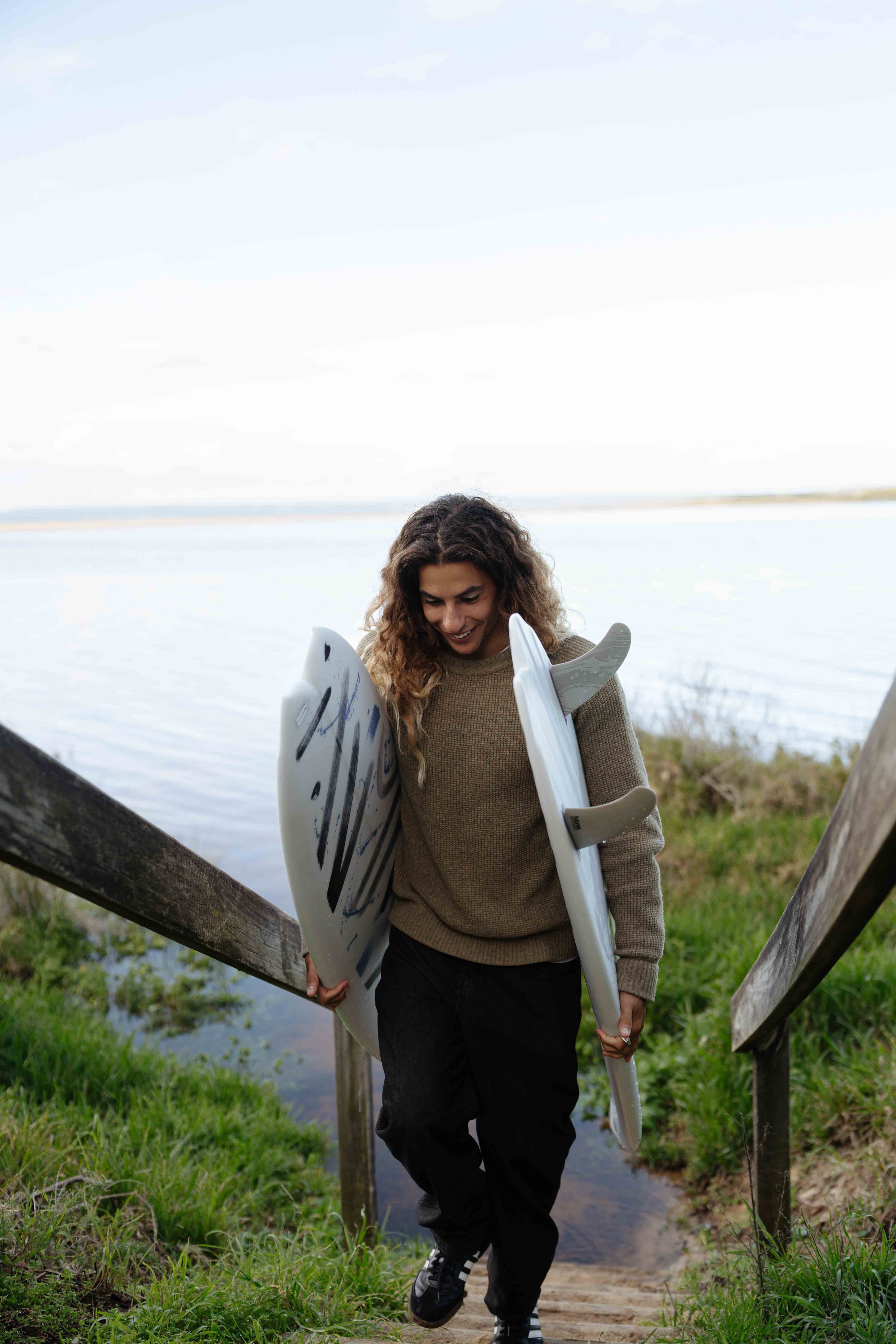 A woman carrying surfboards walking up stairs near a body of water.