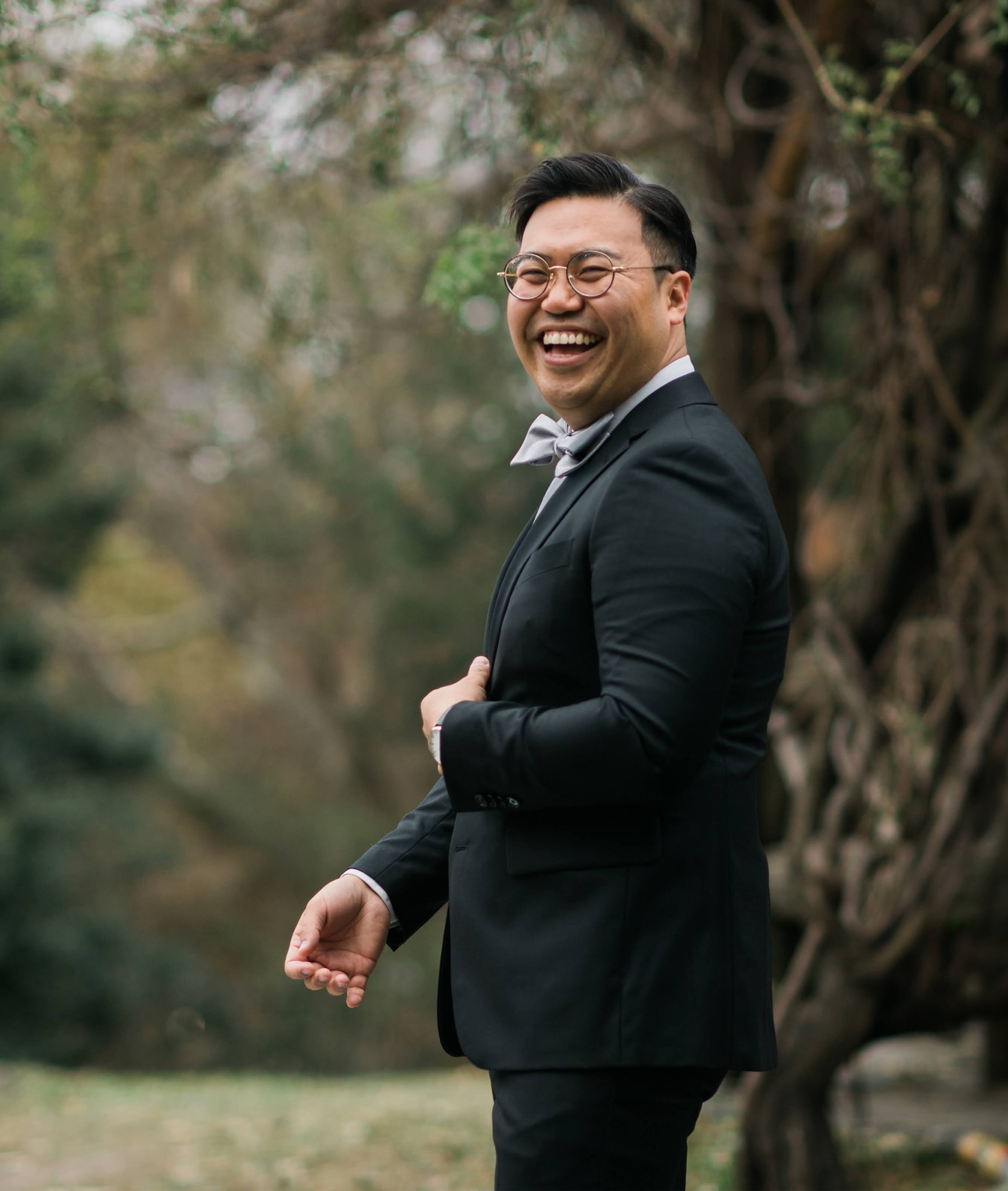A man in a tuxedo with glasses and a bow tie smiling outdoors with trees in the background.