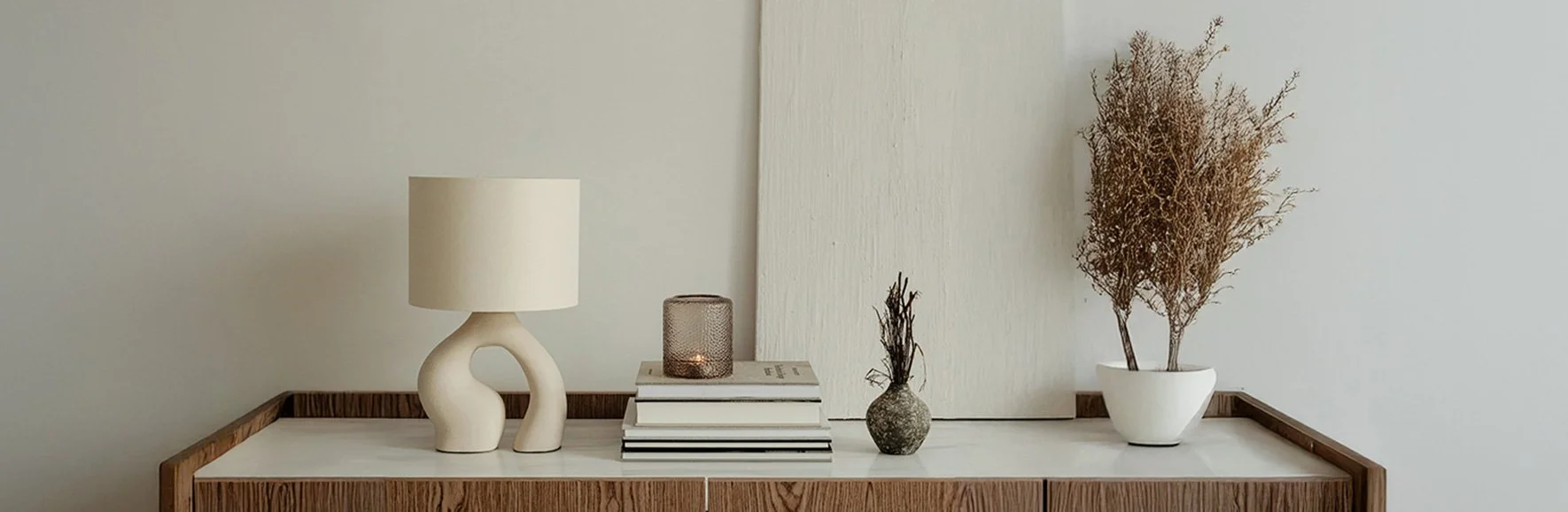 Decorative table with a white lamp, a candle holder, books, a small vase, and a potted dried plant.