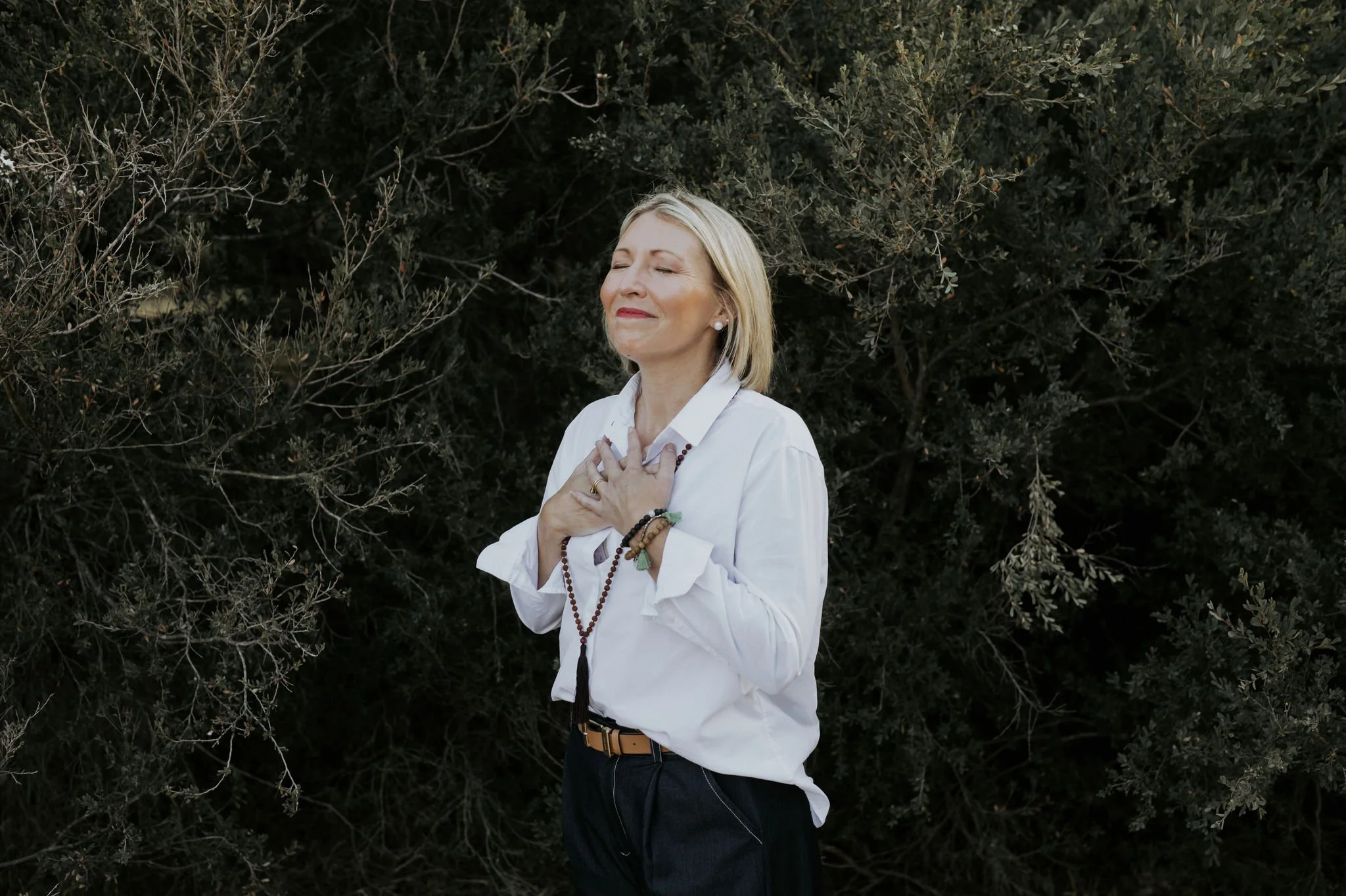A woman with blonde hair wearing a white shirt, spiritual beads, and a bracelet, stands outdoors with her eyes closed and hands over her chest, in front of dark green foliage.
