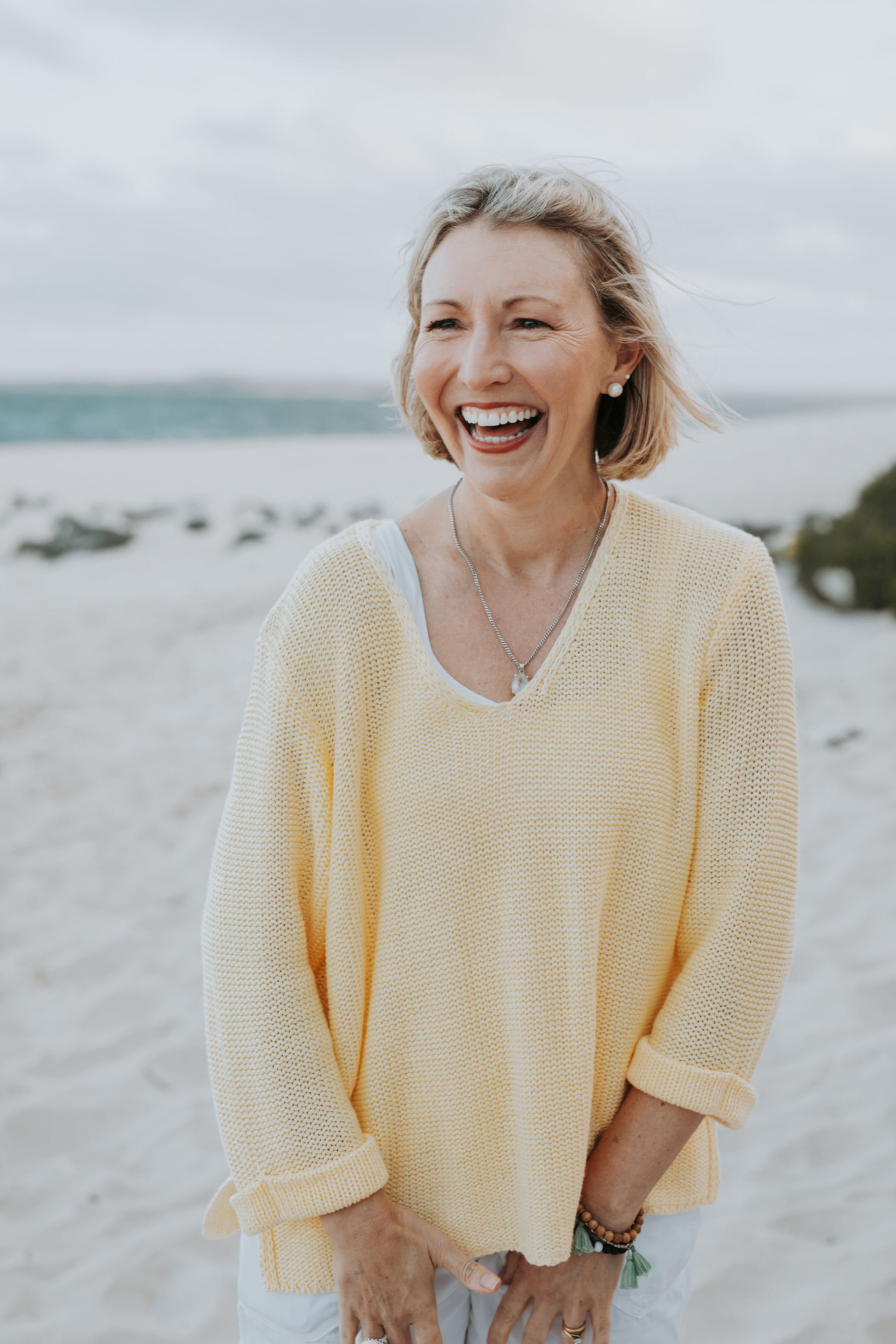 A woman with short blonde hair and a cheerful smile, wearing a yellow sweater, standing on a beach with the ocean and cloudy sky in the background.