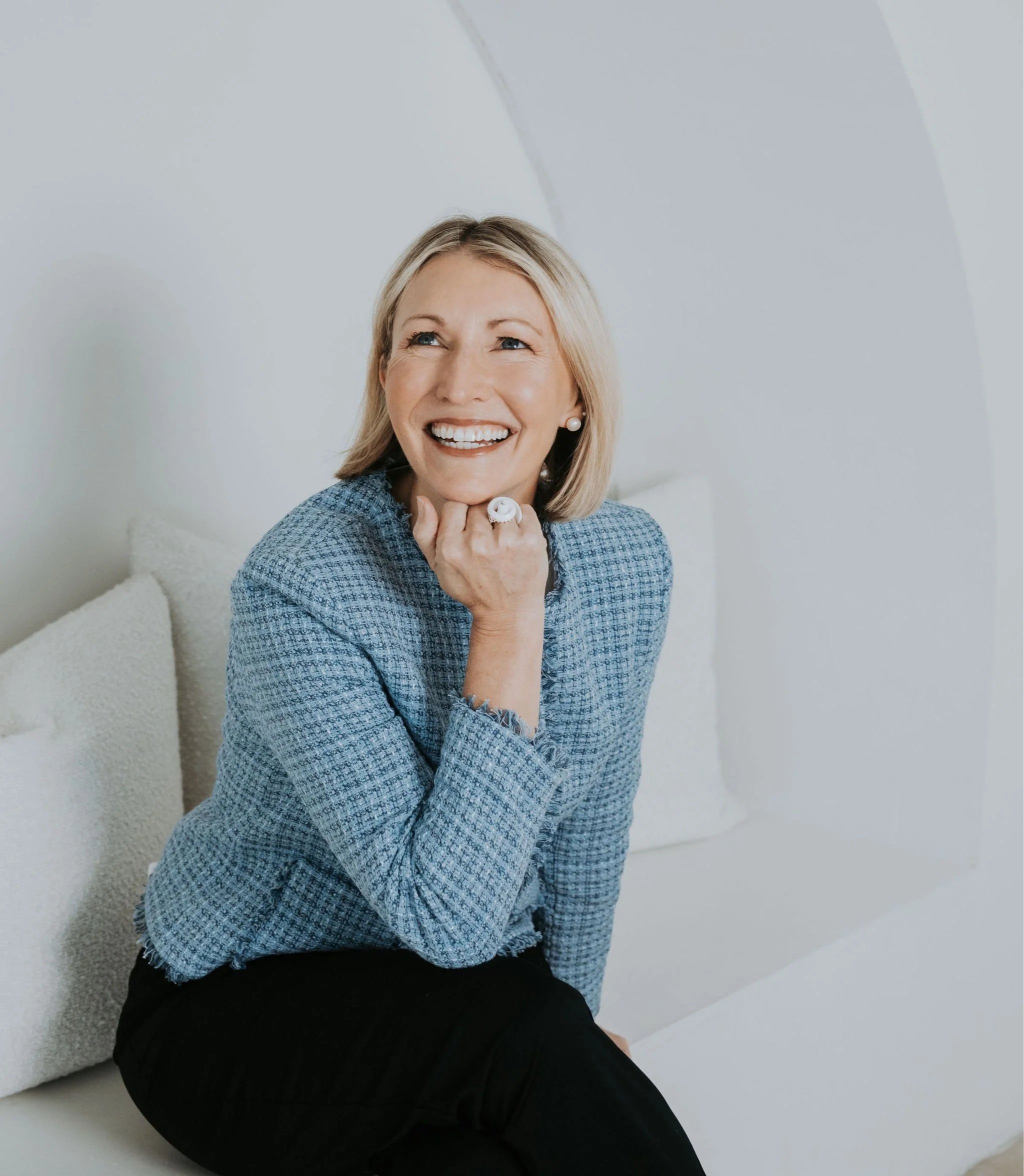 A woman with blonde hair wearing a blue checkered blazer, smiling, sitting on a white sofa in a bright, minimal room.