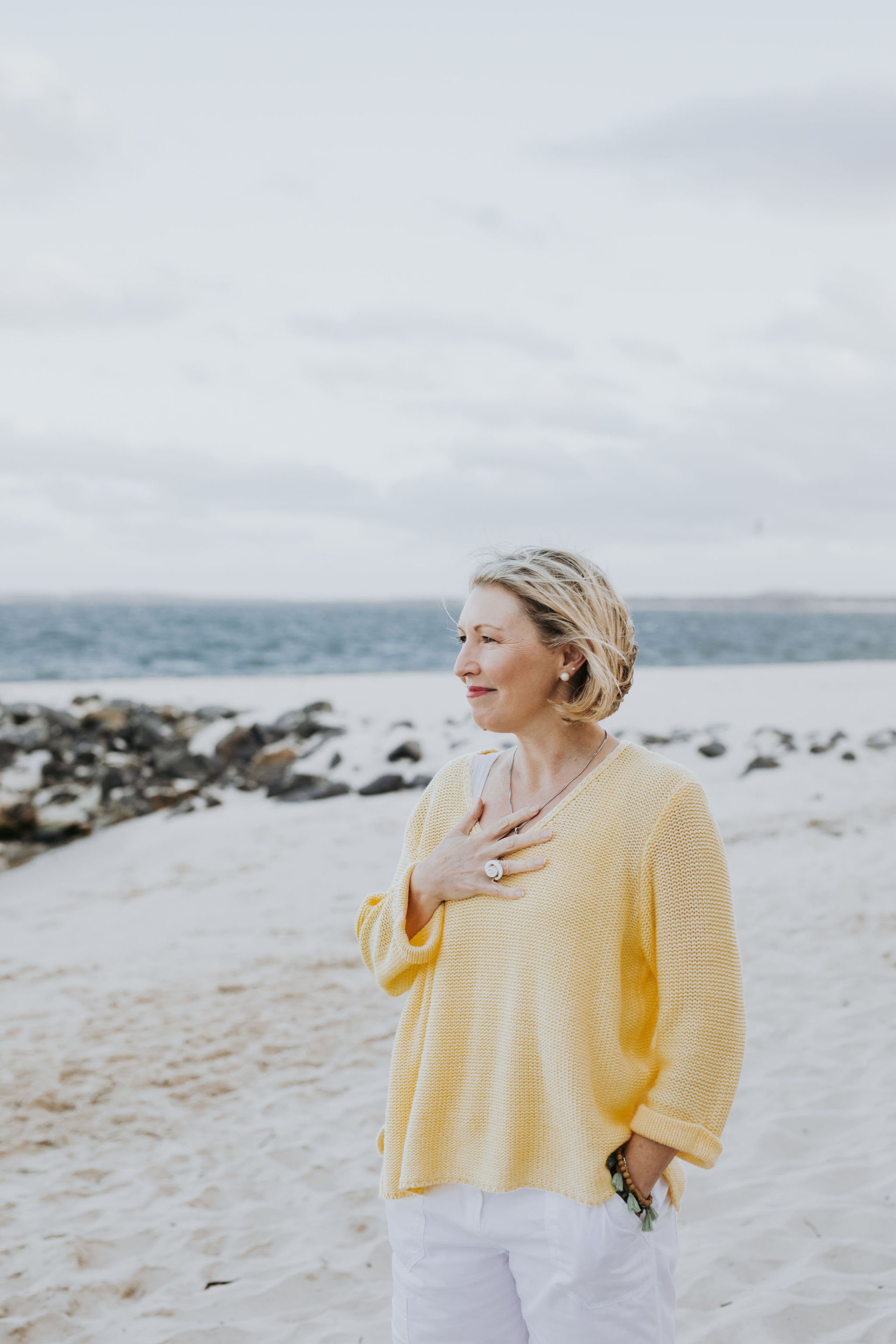 A middle-aged woman standing on a sandy beach with rocks and ocean in the background, wearing a yellow sweater and white pants, with her hand on her chest and looking thoughtful.