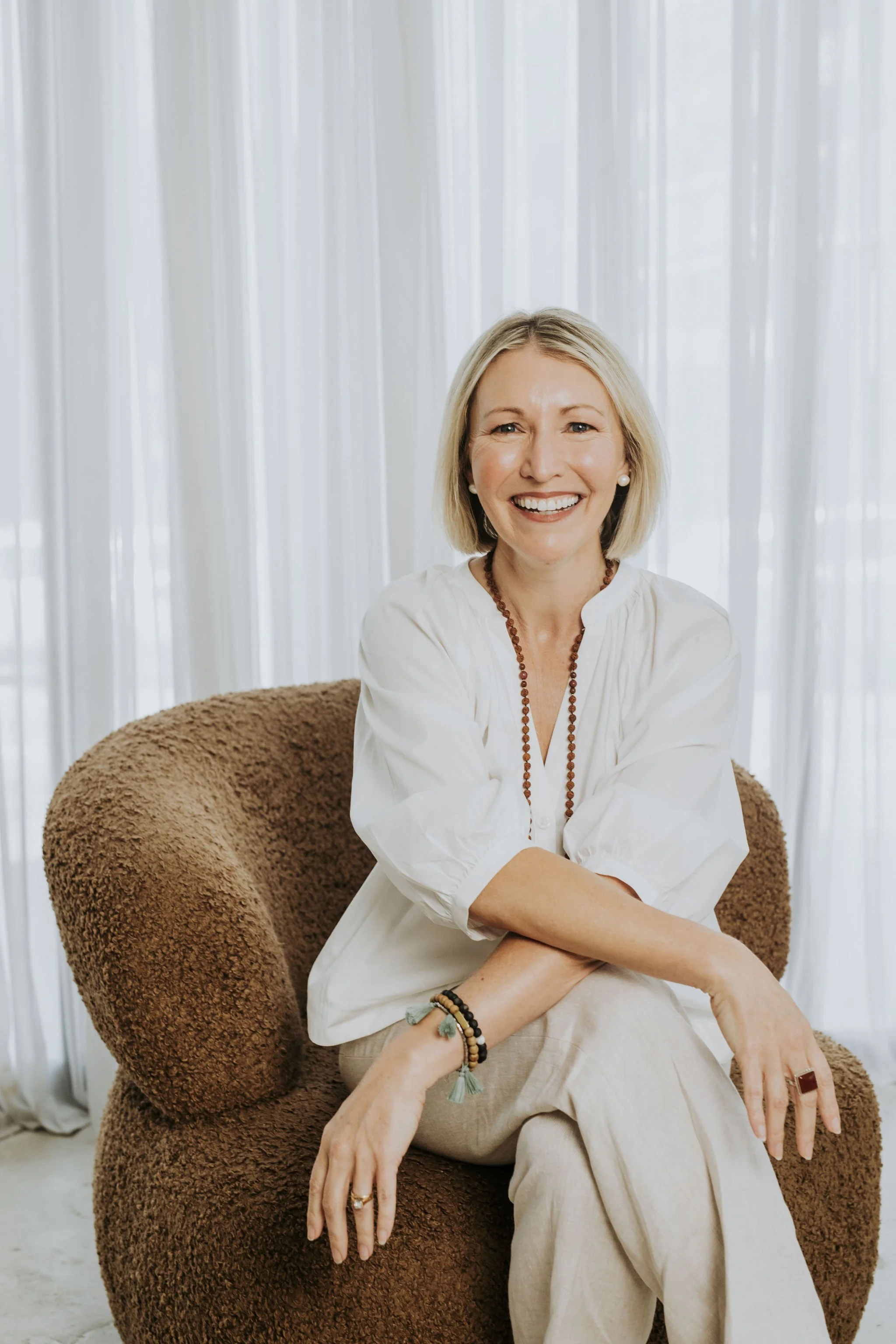 A smiling woman with blonde hair, wearing a white blouse and beige pants, sitting on a brown, textured armchair with a curtain background.