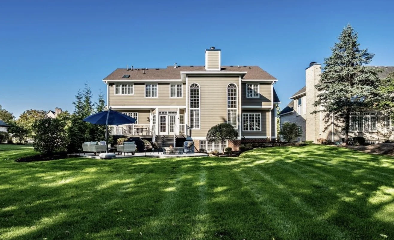 Backyard of a large beige house with multiple windows and a chimney, featuring an outdoor patio with seating, umbrellas, and a well-maintained green lawn under a clear blue sky.