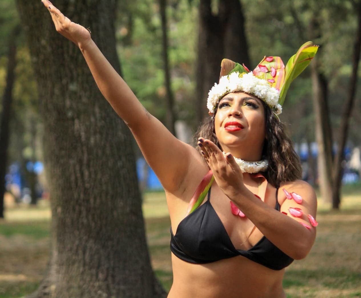 Mujer en un parque durante una ceremonia, vistiendo un disfraz tradicional, con corona de flores y adornos de petalos, realizando un gesto ceremonial.