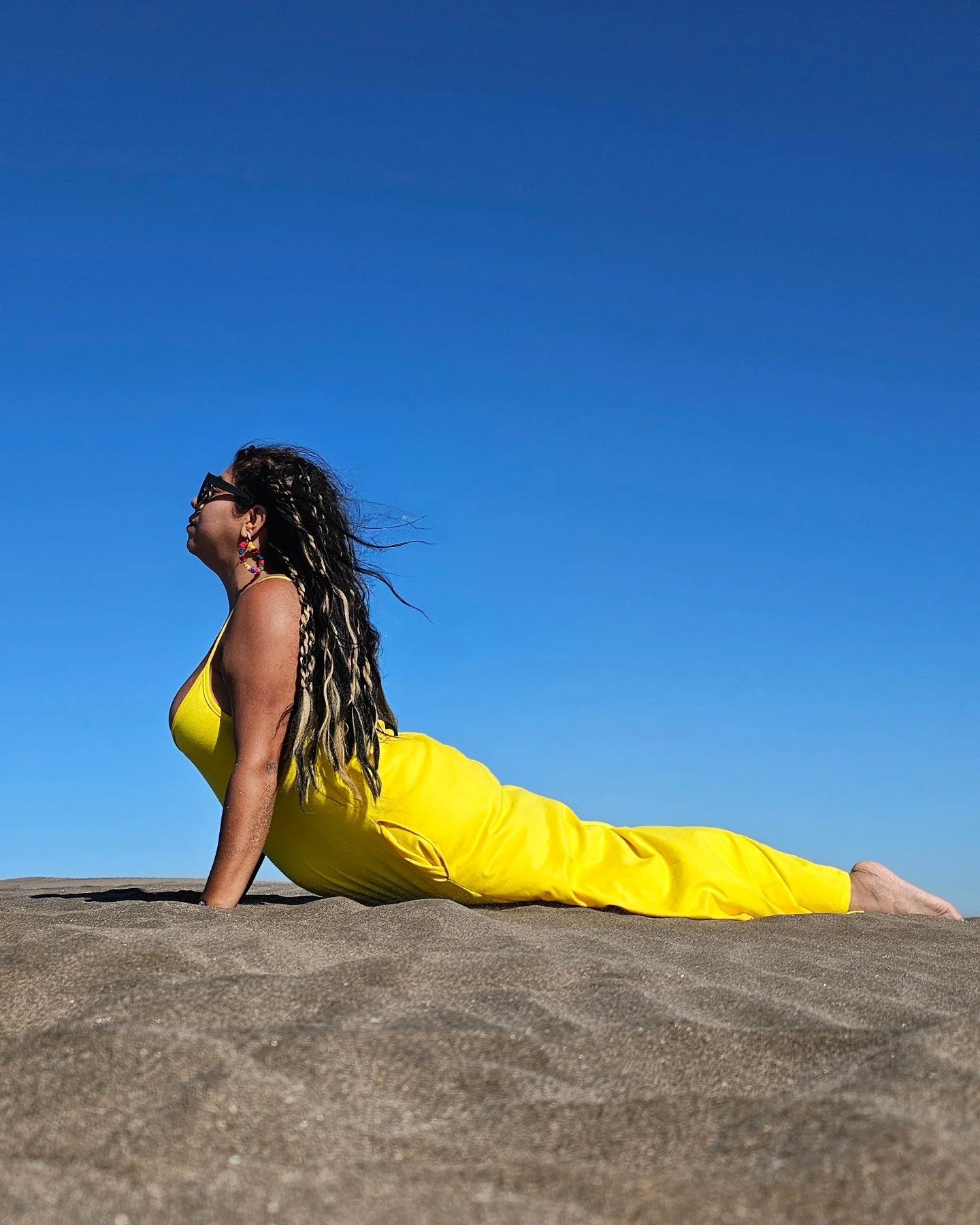 Mujer con cabello rizado, gafas y accesorios coloridos, acostada en arena en posición de yoga, vestida con ropa amarilla, bajo un cielo azul.