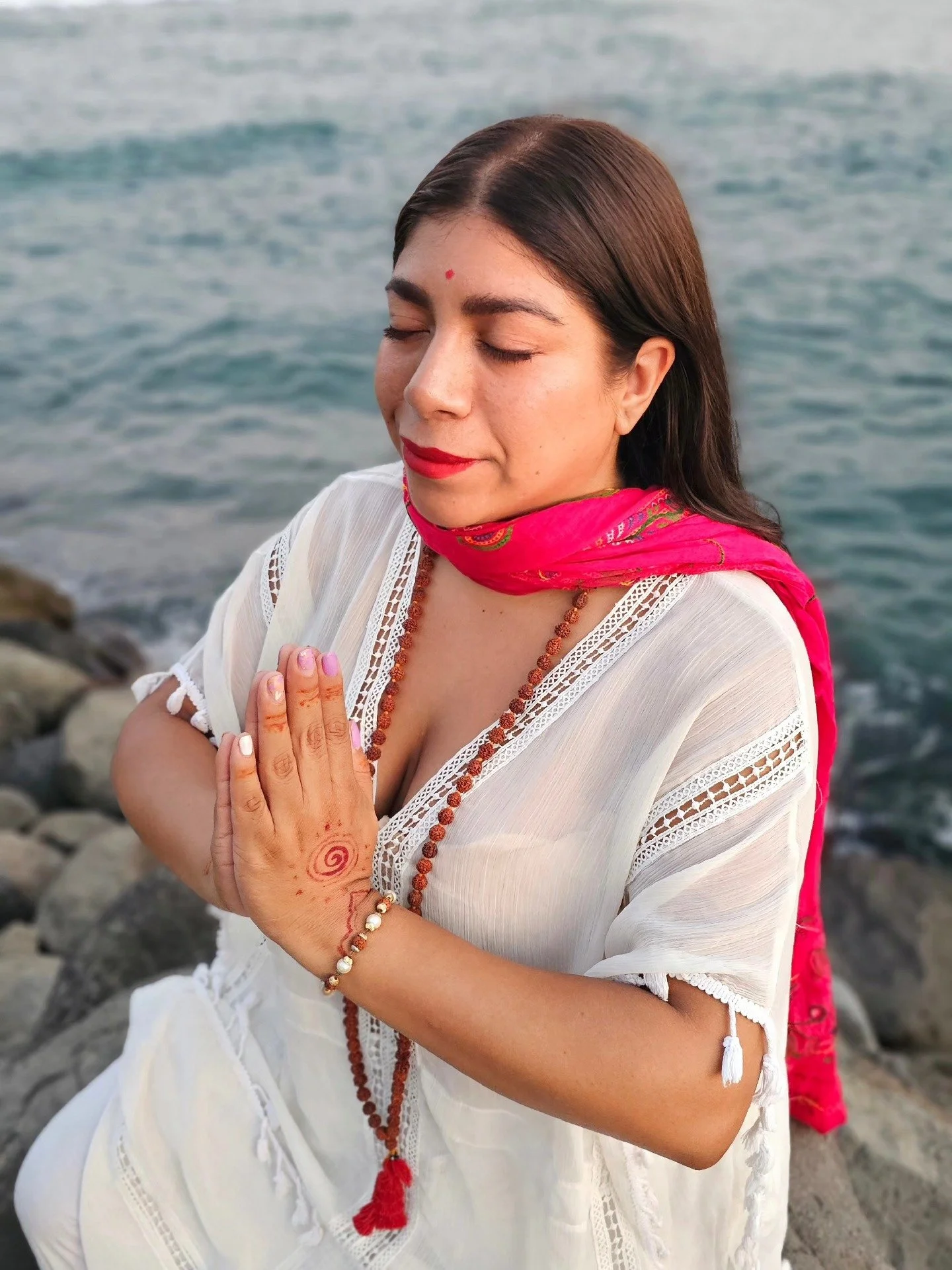 Mujer con los ojos cerrados en una pose de meditación junto al mar, viste ropa blanca, lleva un collar de cuentas y un pañuelo rosa en el cuello, tiene marks en la mano y una recordatoria en el rostro.