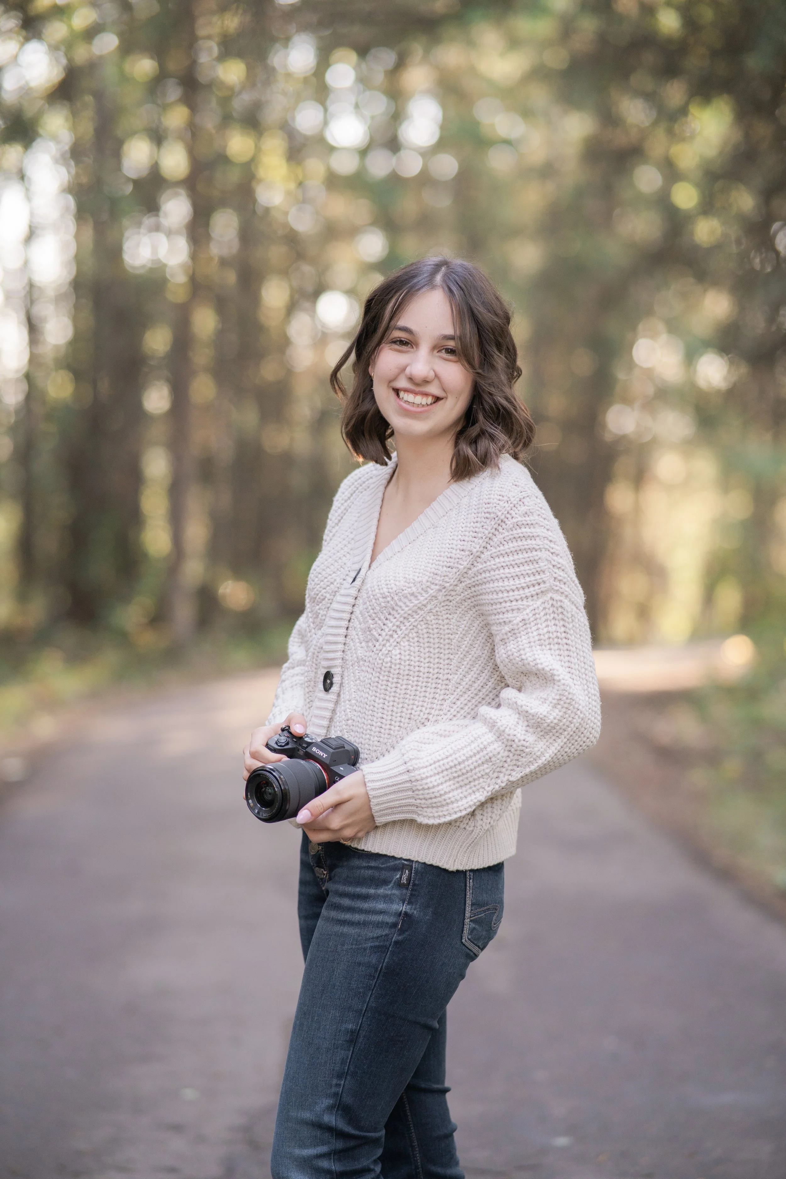 A smiling young woman with shoulder-length brown hair, wearing a cream-colored knitted sweater and jeans, standing on a forest path holding a camera.