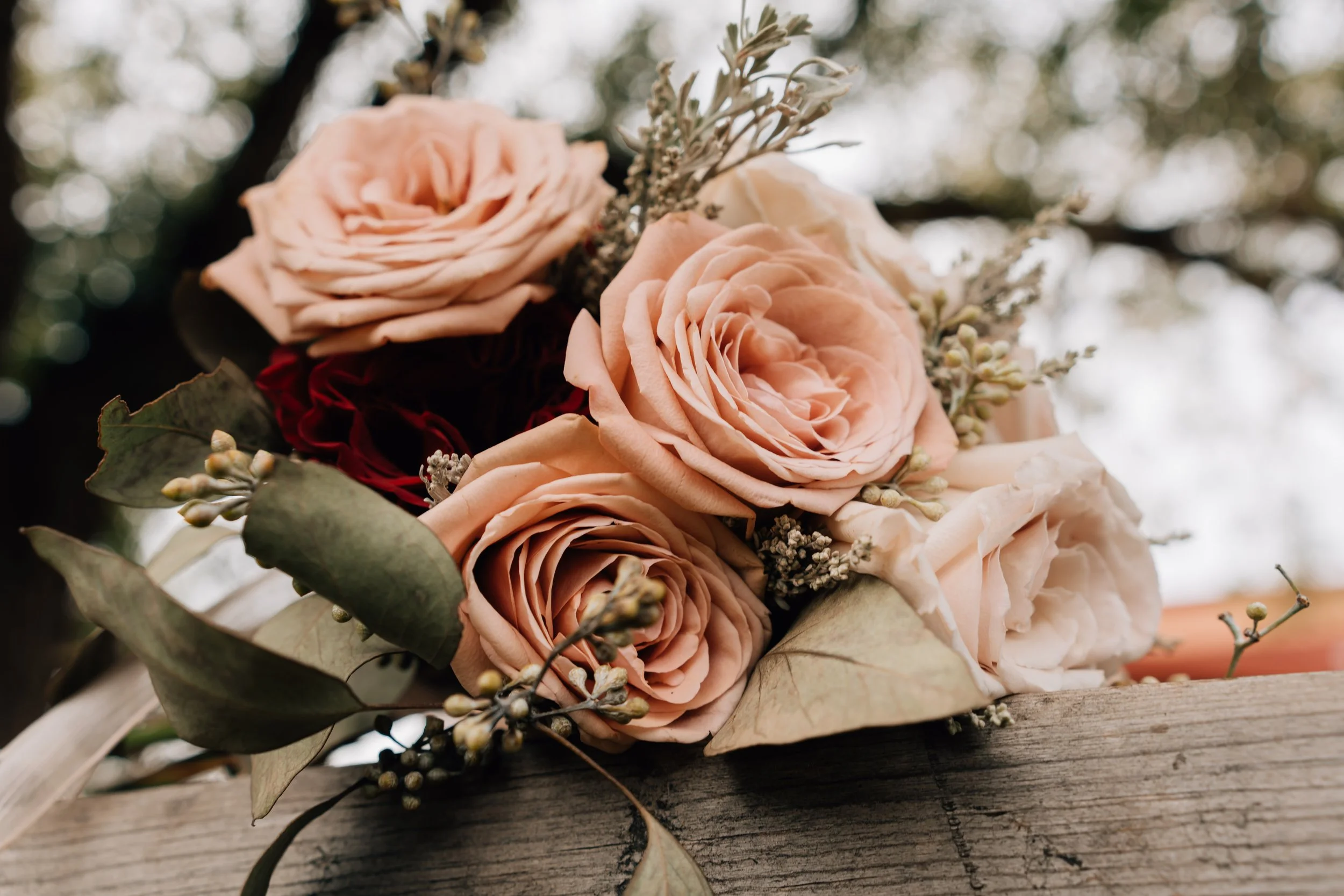 Close-up of a bouquet of light pink roses, greenery, and small white flowers on a rustic wooden surface against a blurred outdoor background.
