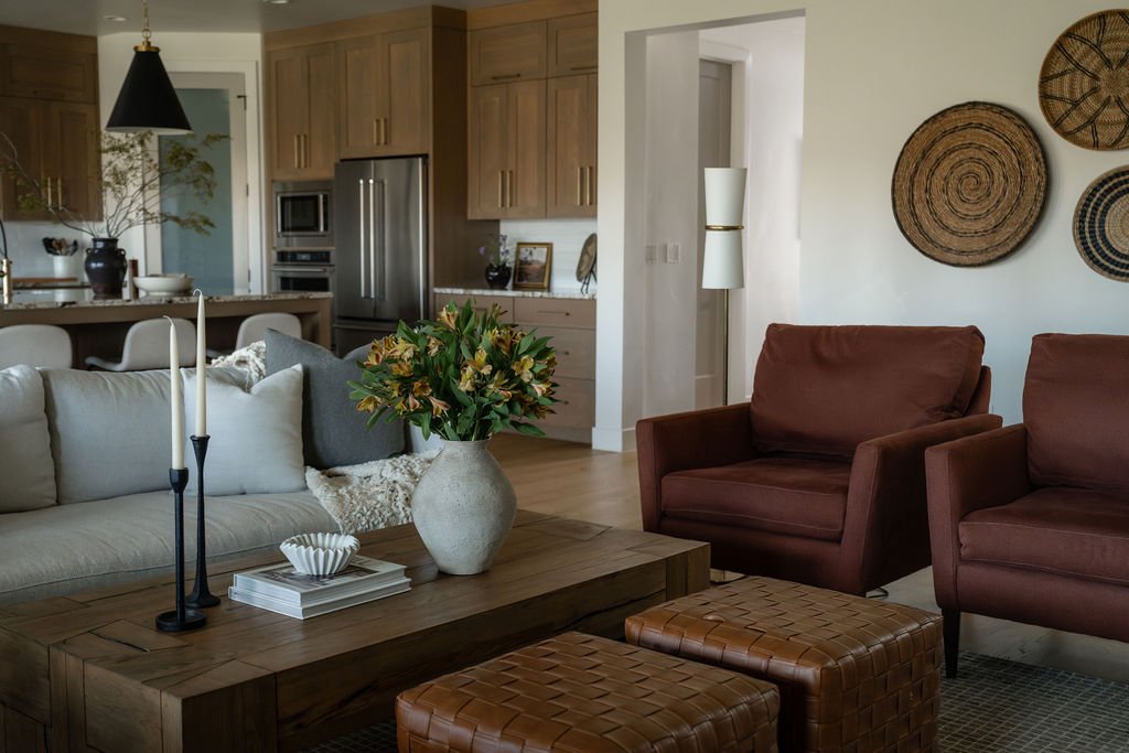 Living room with beige sofa, two brown armchairs, a wooden coffee table with a vase of flowers, and wall decor. Kitchen in background with cabinets and stainless steel appliances.