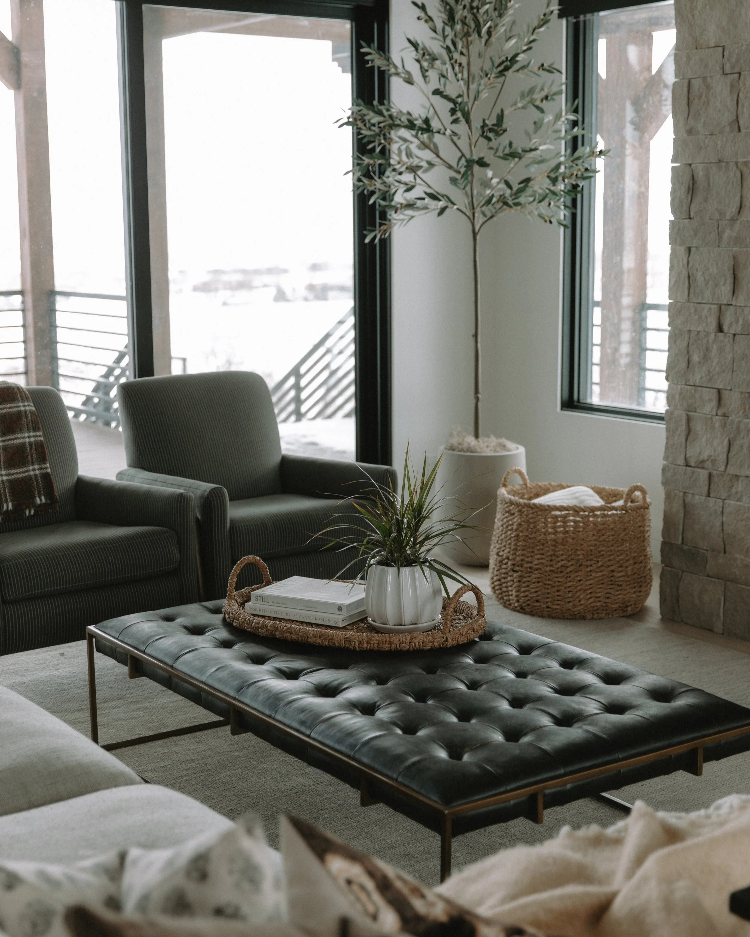 Cozy living room with neutral furniture, a black leather ottoman, soft textiles, and large windows creating a bright and inviting space.
