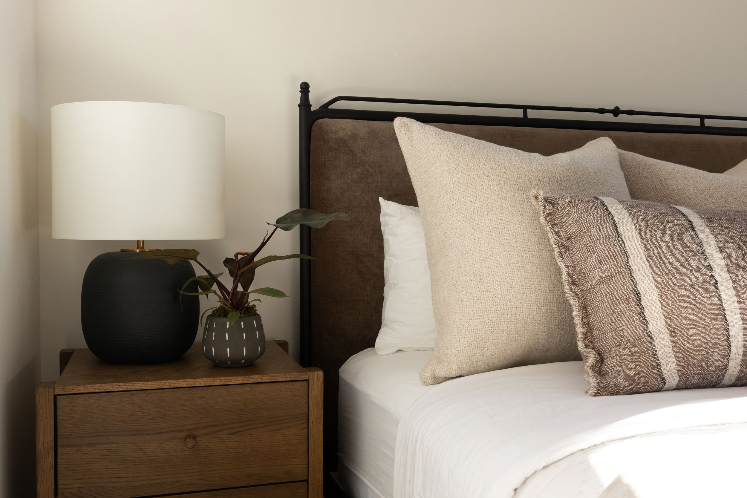 A bedroom nightstand with a white lampshade and a potted plant, next to a bed with beige pillows and a brown headboard.