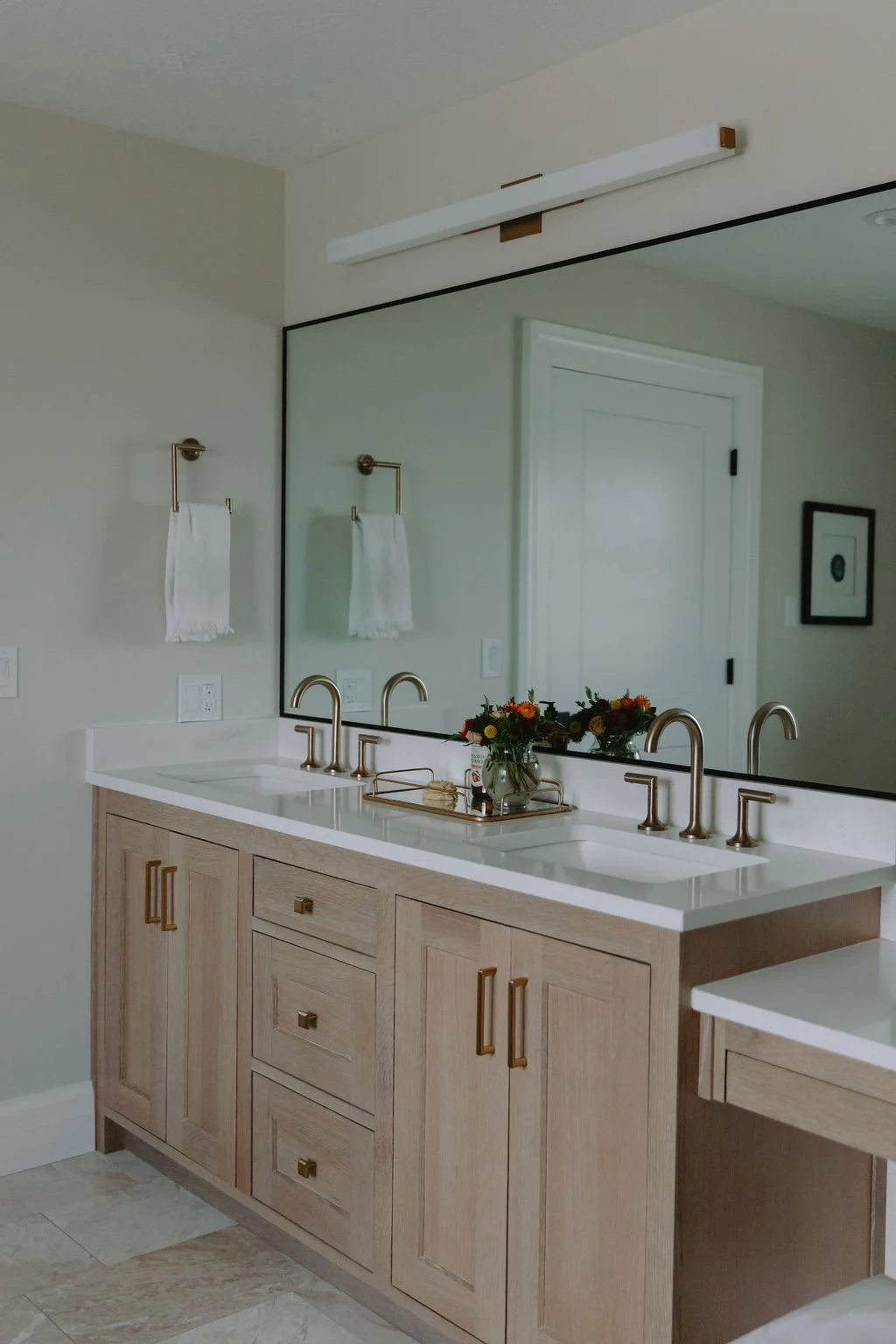 Elegant double vanity with light wood cabinetry, gold faucets, a large mirror, and fresh floral styling.