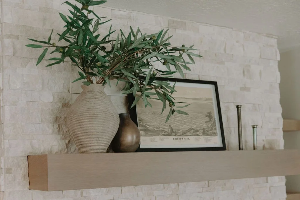 Close-up of a styled fireplace mantel with textured stone, a framed landscape print, and organic ceramic vases.