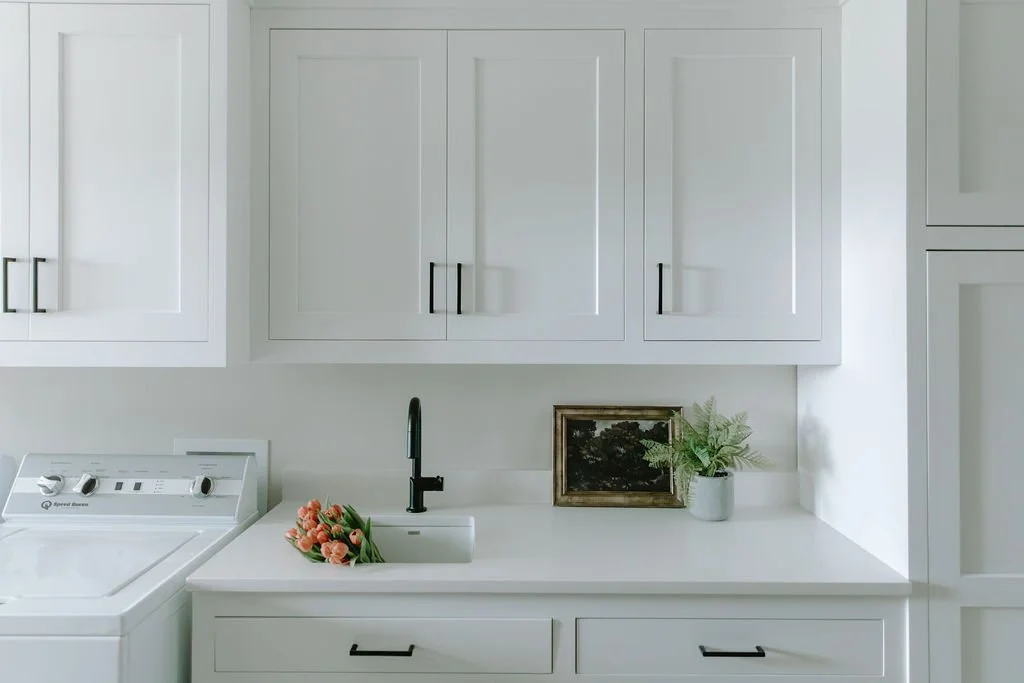 Minimalist laundry area with white cabinetry, a built-in sink, and fresh greenery adding warmth to the space.