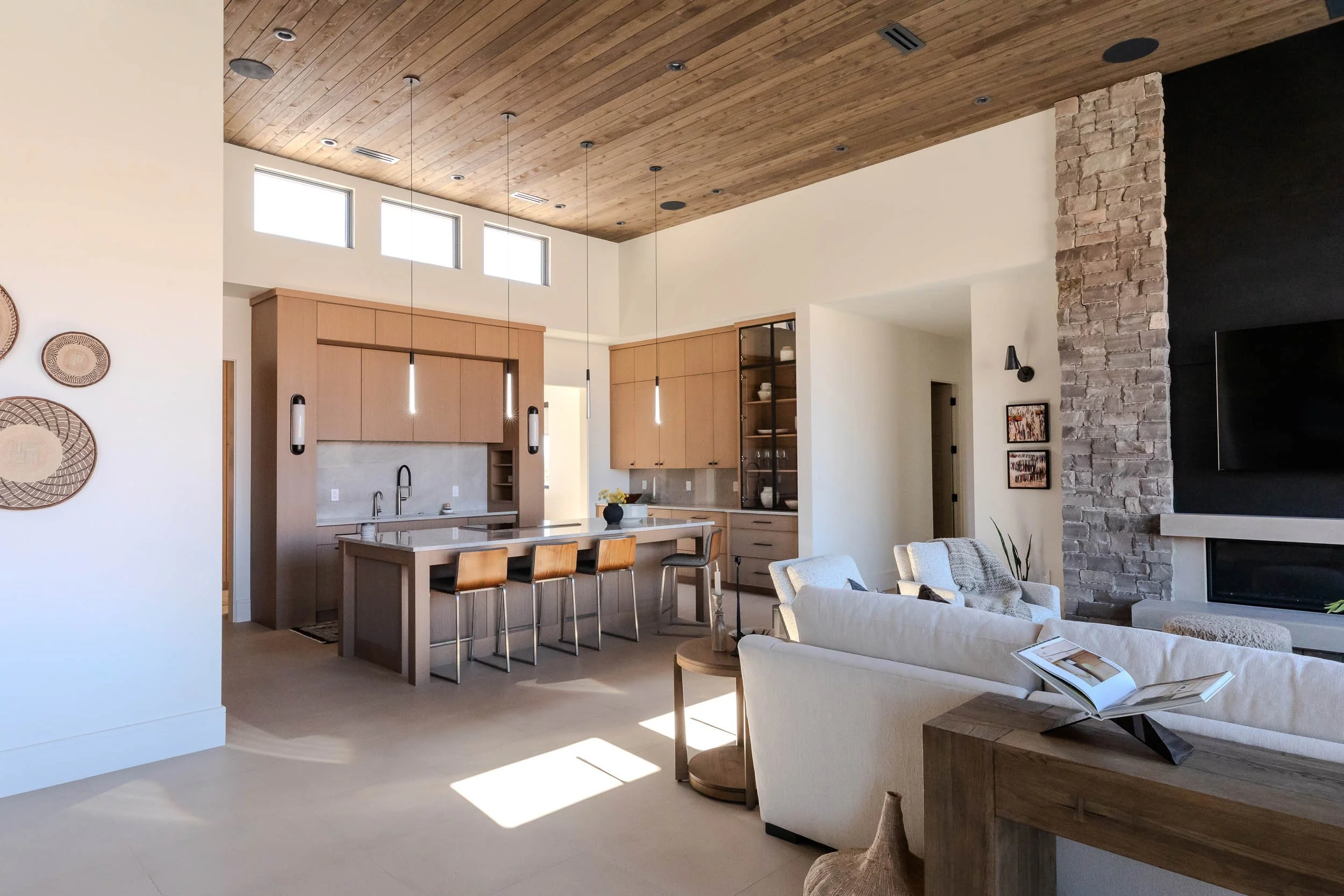 Open-concept living room and kitchen with high wooden ceiling, beige furniture, stone fireplace, and natural light from windows.