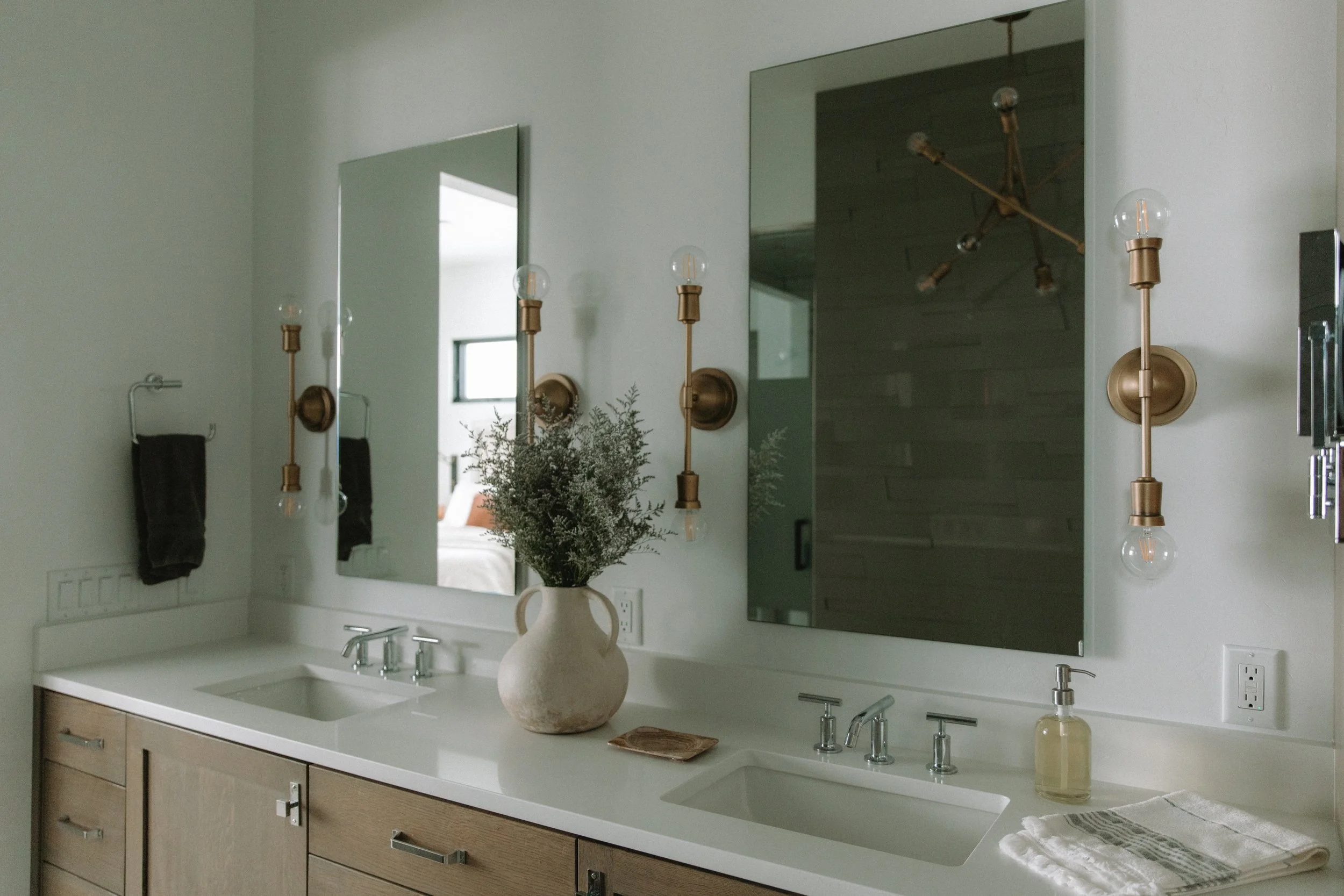 Modern master bathroom with double sinks, gold wall lighting, and greenery in vase