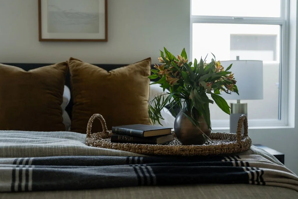 Cozy bedroom setting with a woven tray, fresh greenery, and layered textiles on a plush bed with warm-toned accent pillows.