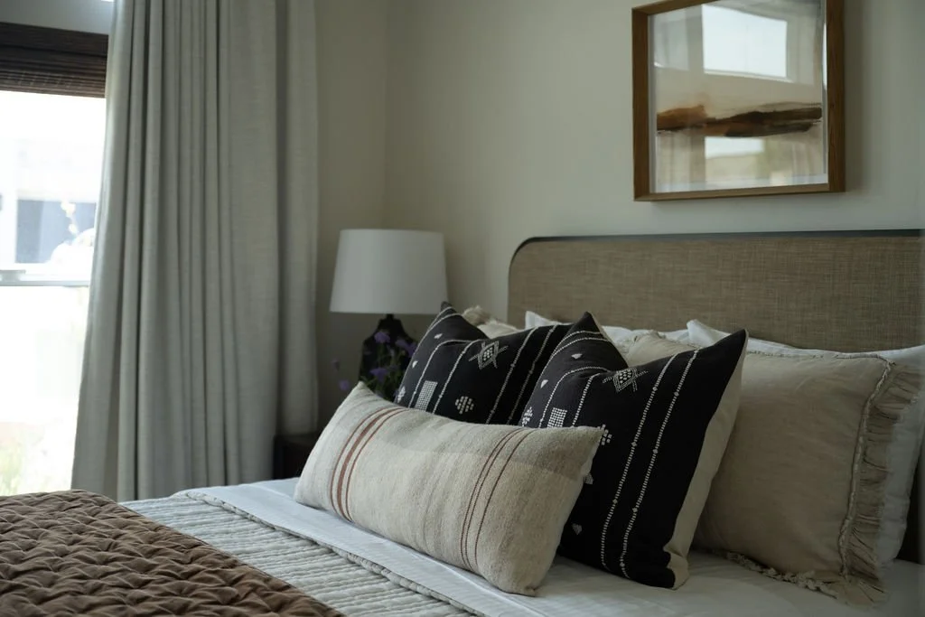 Close-up of a cozy bed with textured throw pillows, layered neutral bedding, and warm wood nightstand styling.
