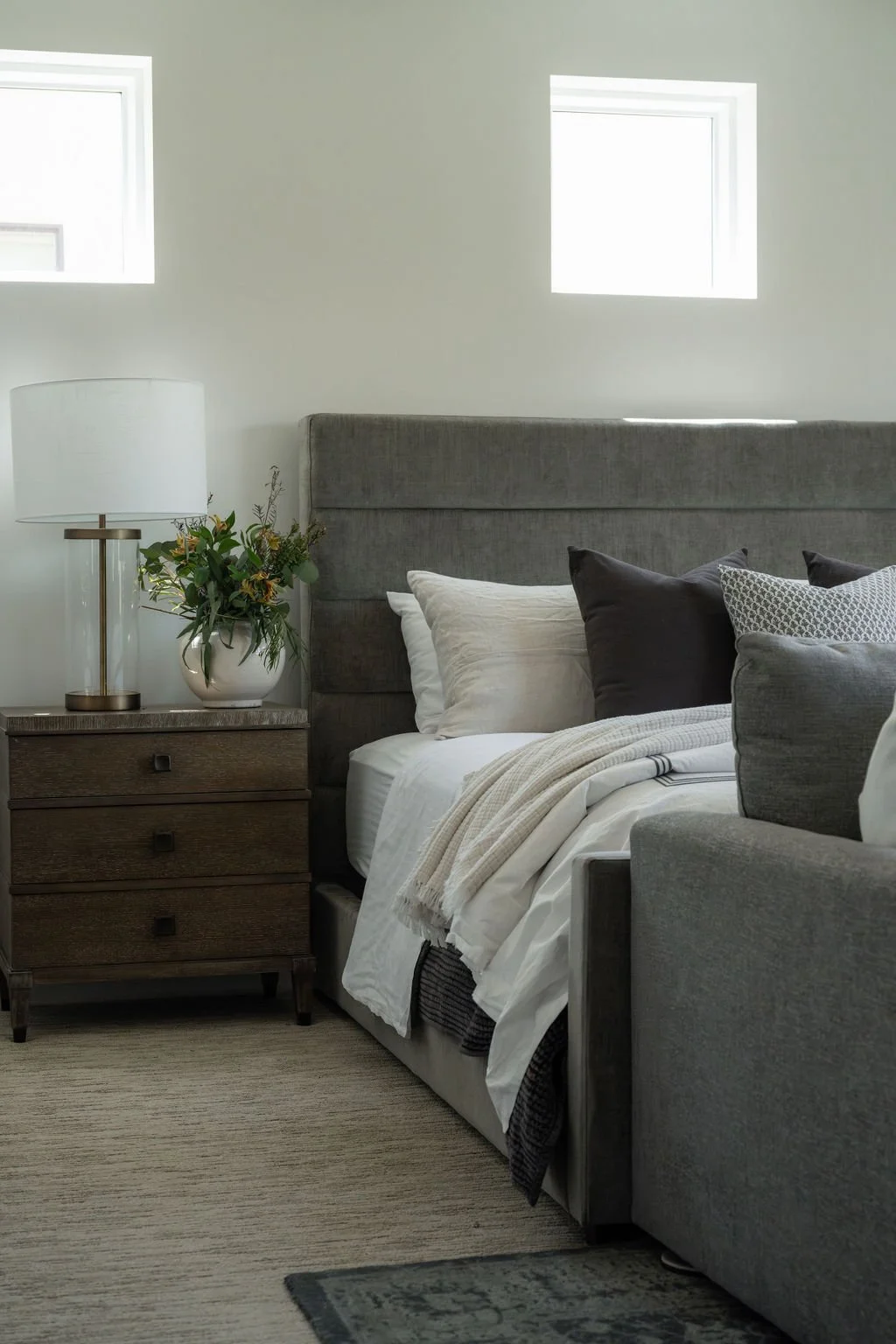 Serene bedroom with a gray upholstered bed, layered bedding, and a wood nightstand with a glass lamp and fresh greenery.
