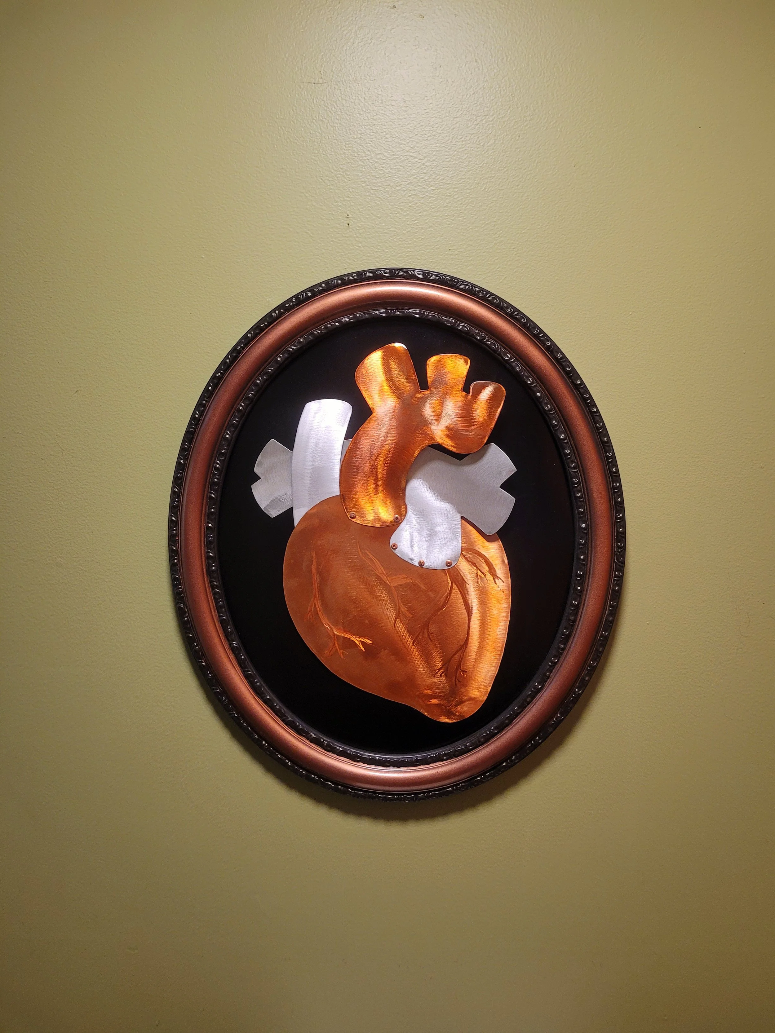 Artwork of a human heart made of metallic materials, framed and mounted on a green wall.