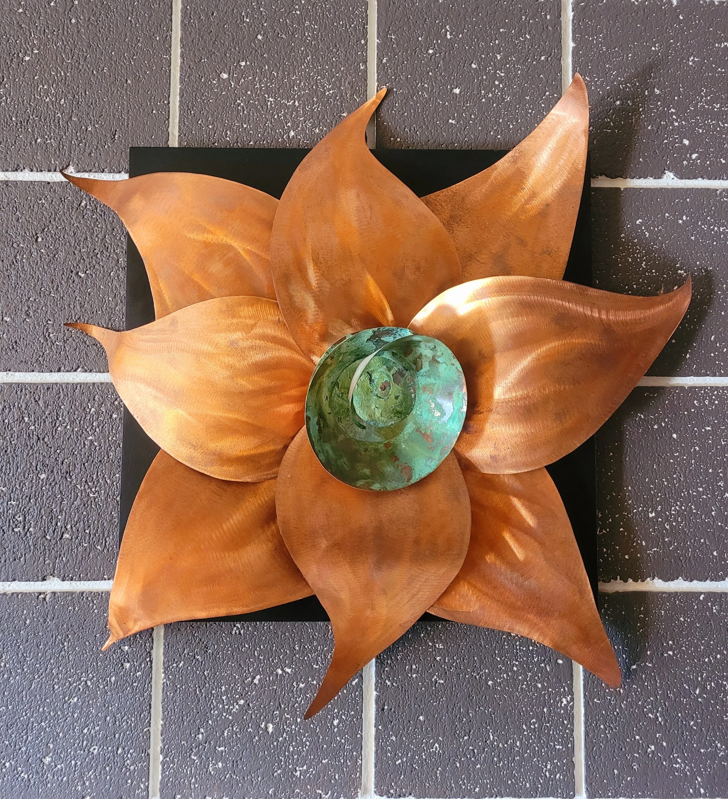 Decorative wall sculpture resembling a flower with large copper-colored metal petals and a central green glass element, mounted on a black square background, against a tiled wall.
