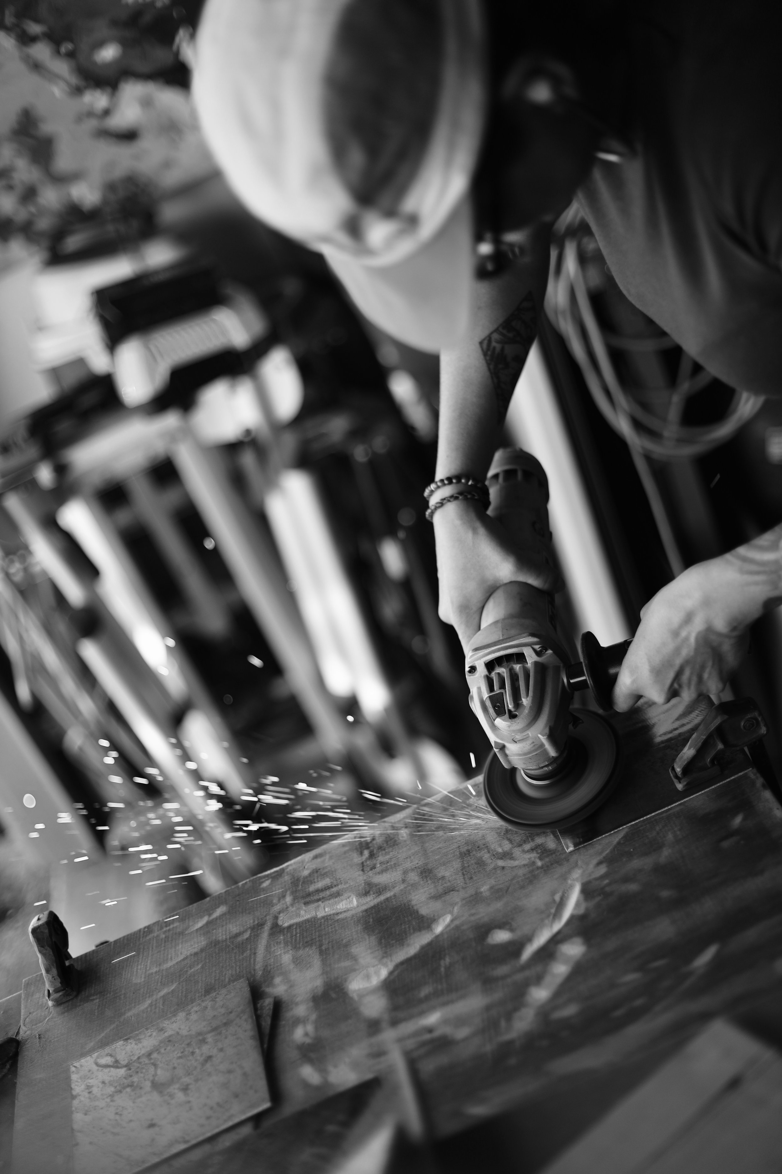 A person wearing a cap and bracelet uses an angle grinder on a wooden surface, creating sparks, in a workshop or carpentry environment.