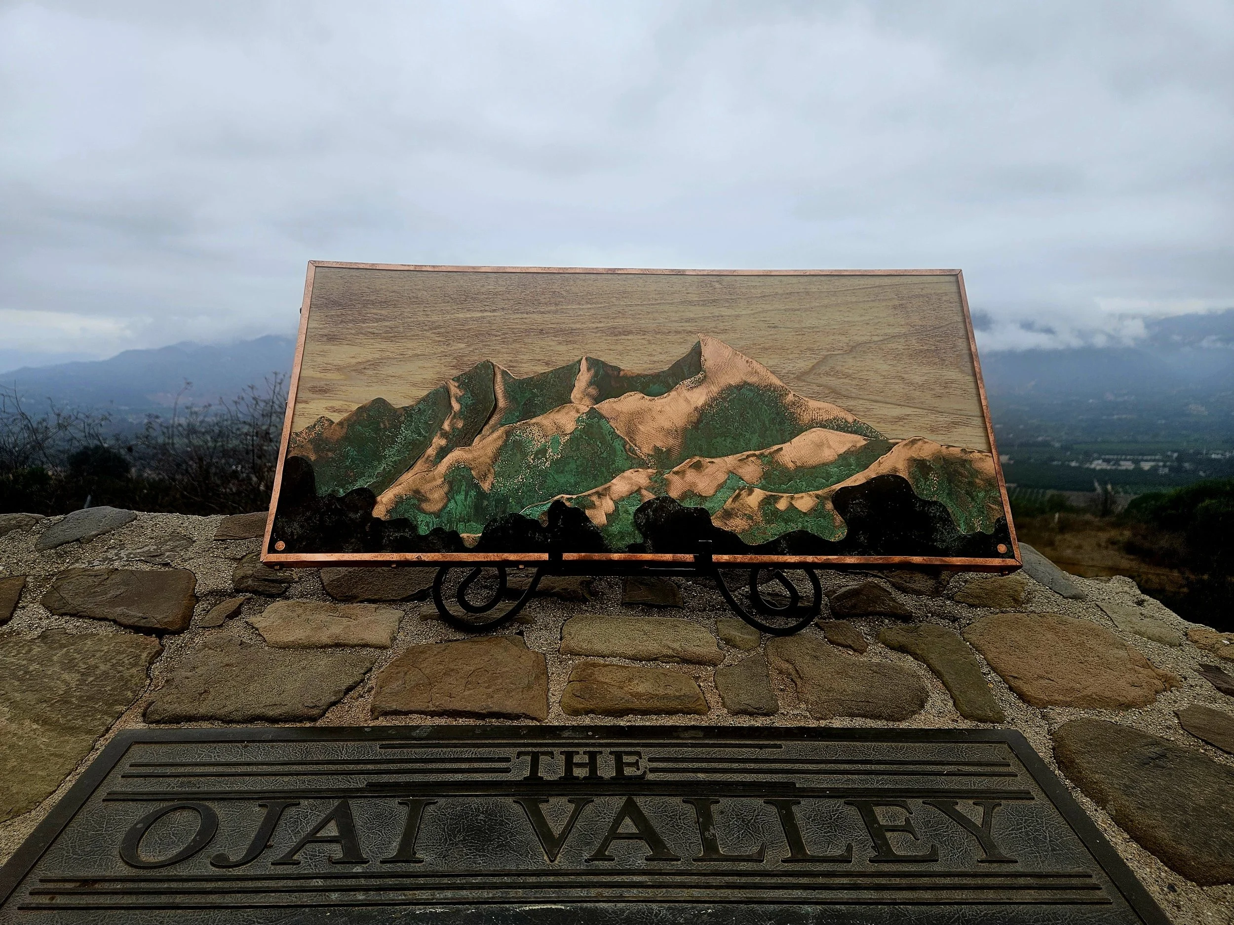 A sign reading 'The Ojai Valley' and a painting of mountains on a wooden board, placed on a stone wall with an outdoor landscape in the background.