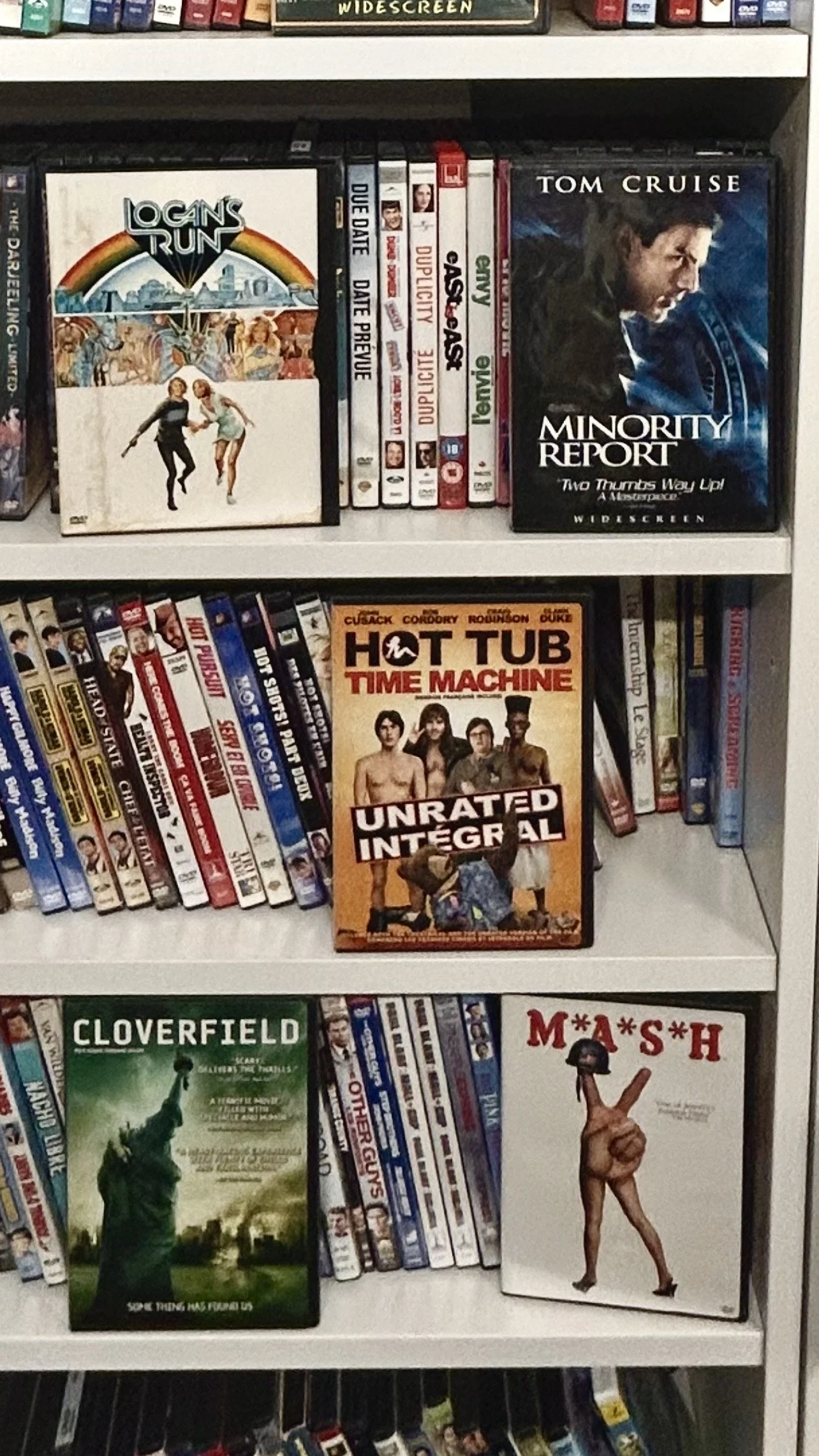 Shelf containing DVDs and books, including movies like 'Logan's Run,' 'Minority Report,' 'Hot Tub Time Machine,' 'Cloverfield,' and 'M*A*S*H'.