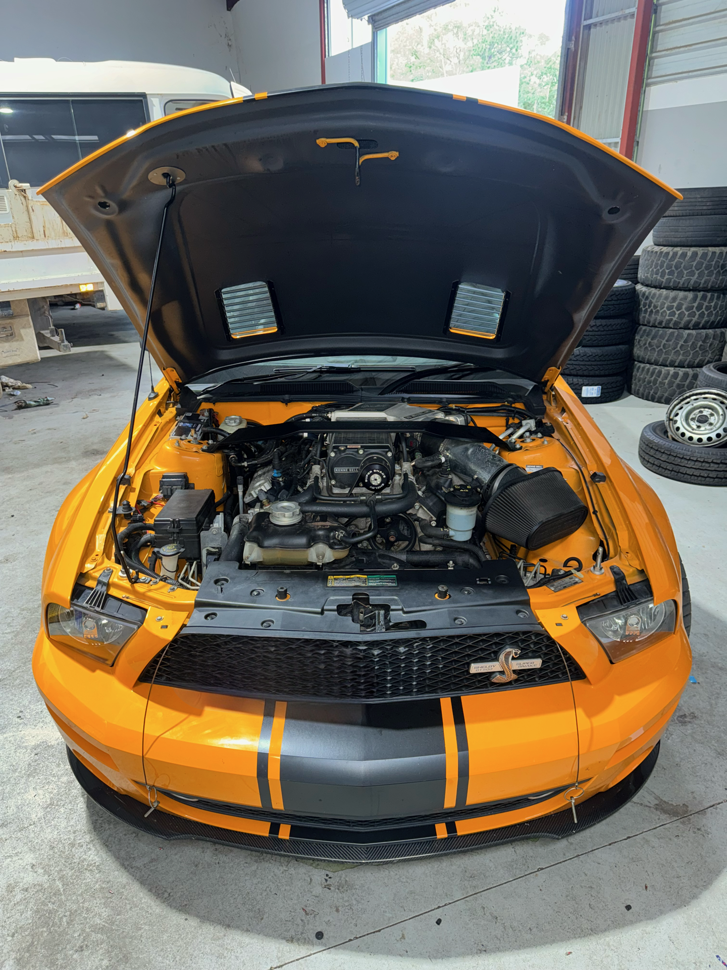 Yellow Ford Mustang with black racing stripes, hood open showing engine bay inside a garage with spare tires on the right side.