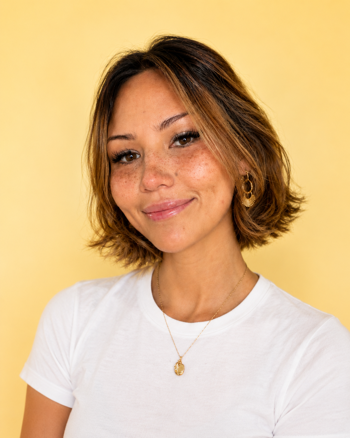 A woman with short, wavy brown hair with lighter highlights, smiling, wearing a white t-shirt, gold earrings, and a gold necklace with a pendant, against a yellow background.