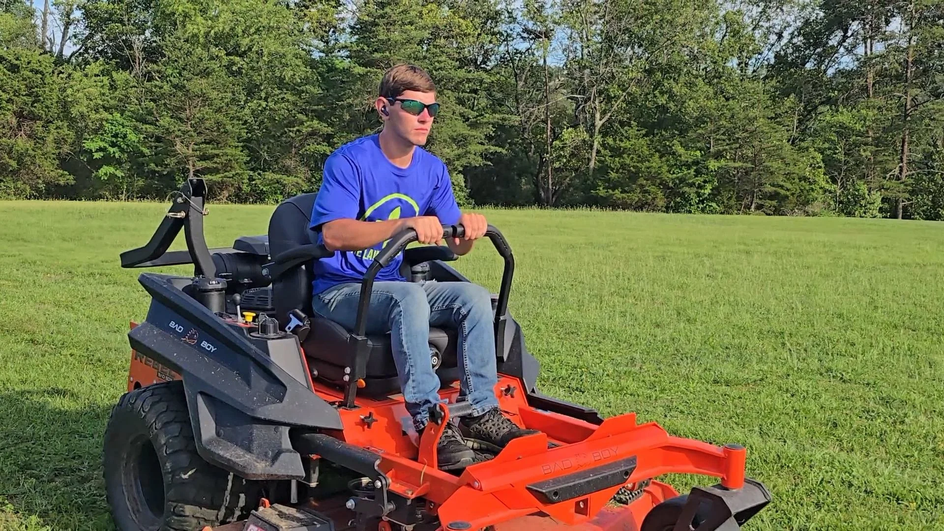 A young man wearing sunglasses and a blue Lawn Locals T-shirt operating a riding mower on a lawn with trees in the background.