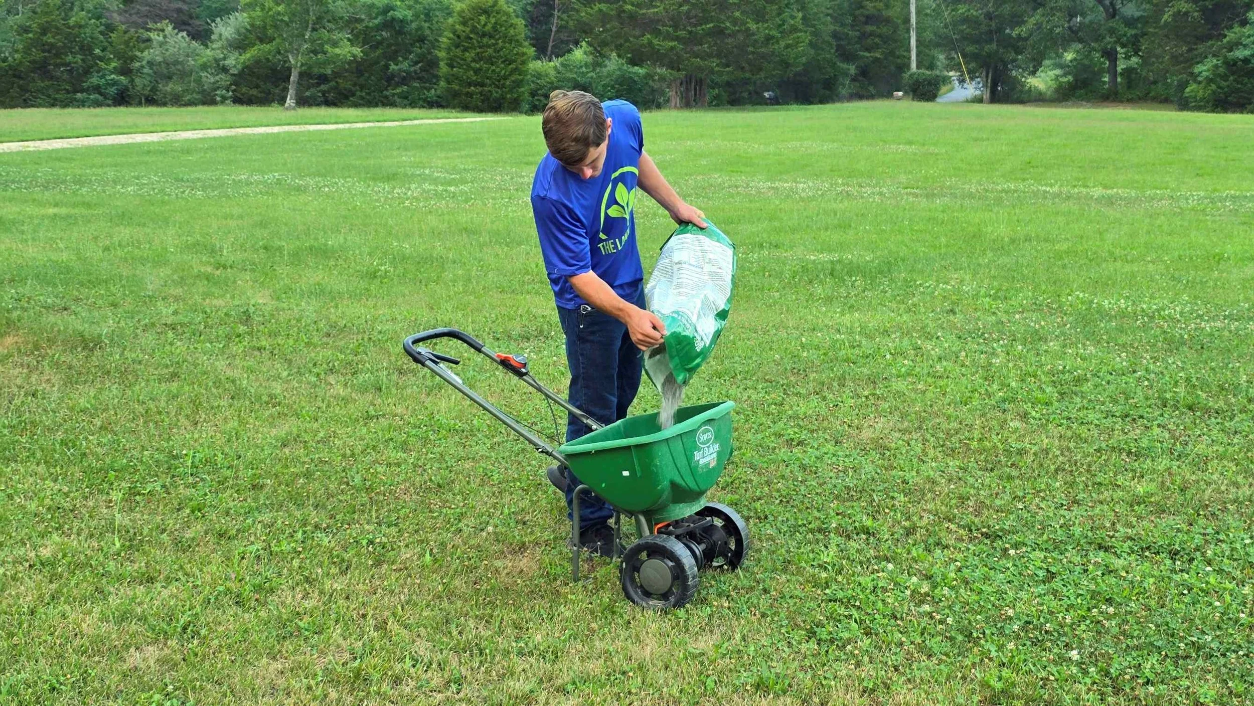 A young man wearing a blue Lawn Locals T-shirt and blue jeans is pouring fertilizer or seed from a bag into a small green broadcast spreader in a grassy field, with trees and a gravel driveway in the background.