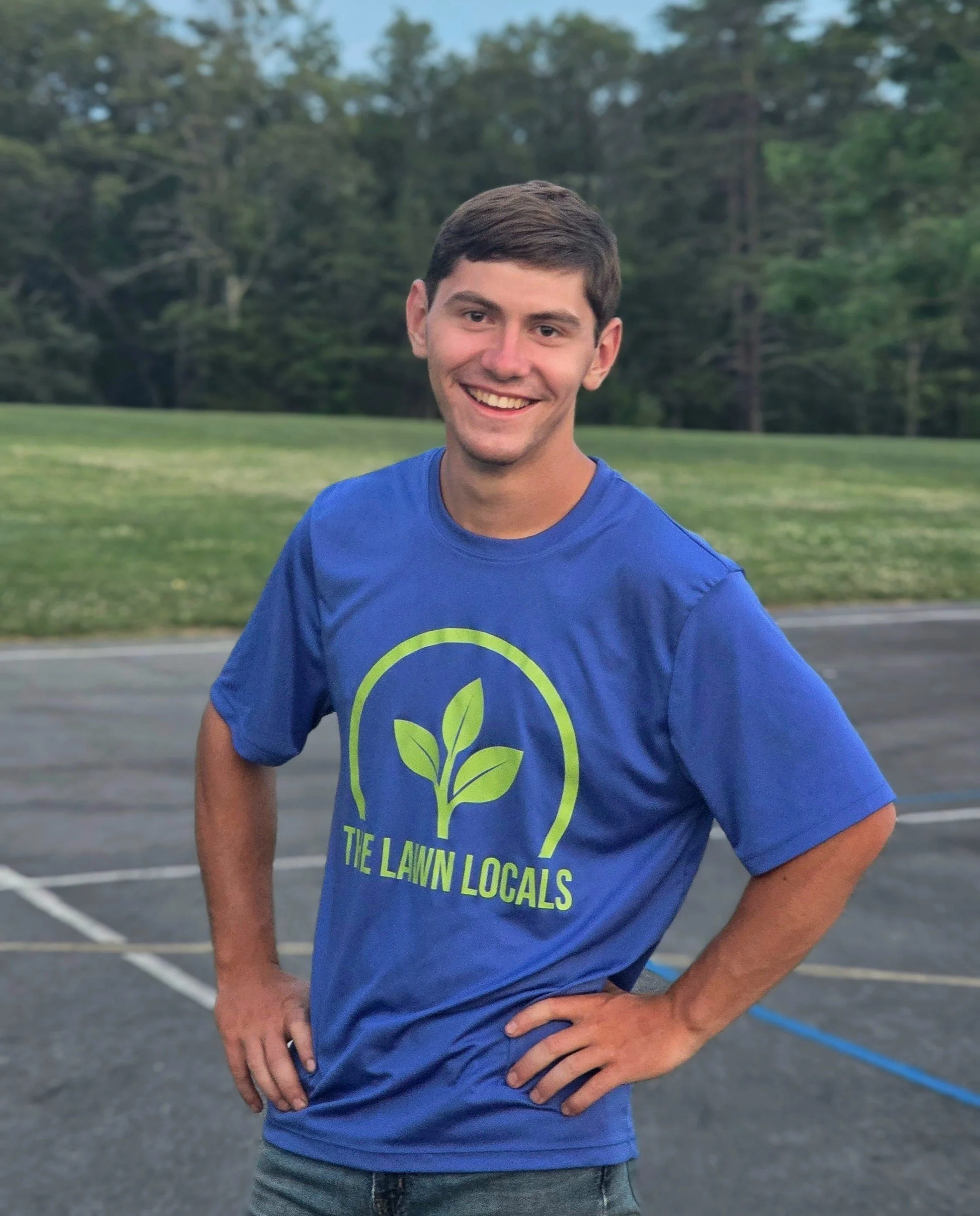 A young man smiling outdoors, wearing a blue Lawn Locals T-shirt, standing with trees in the background.