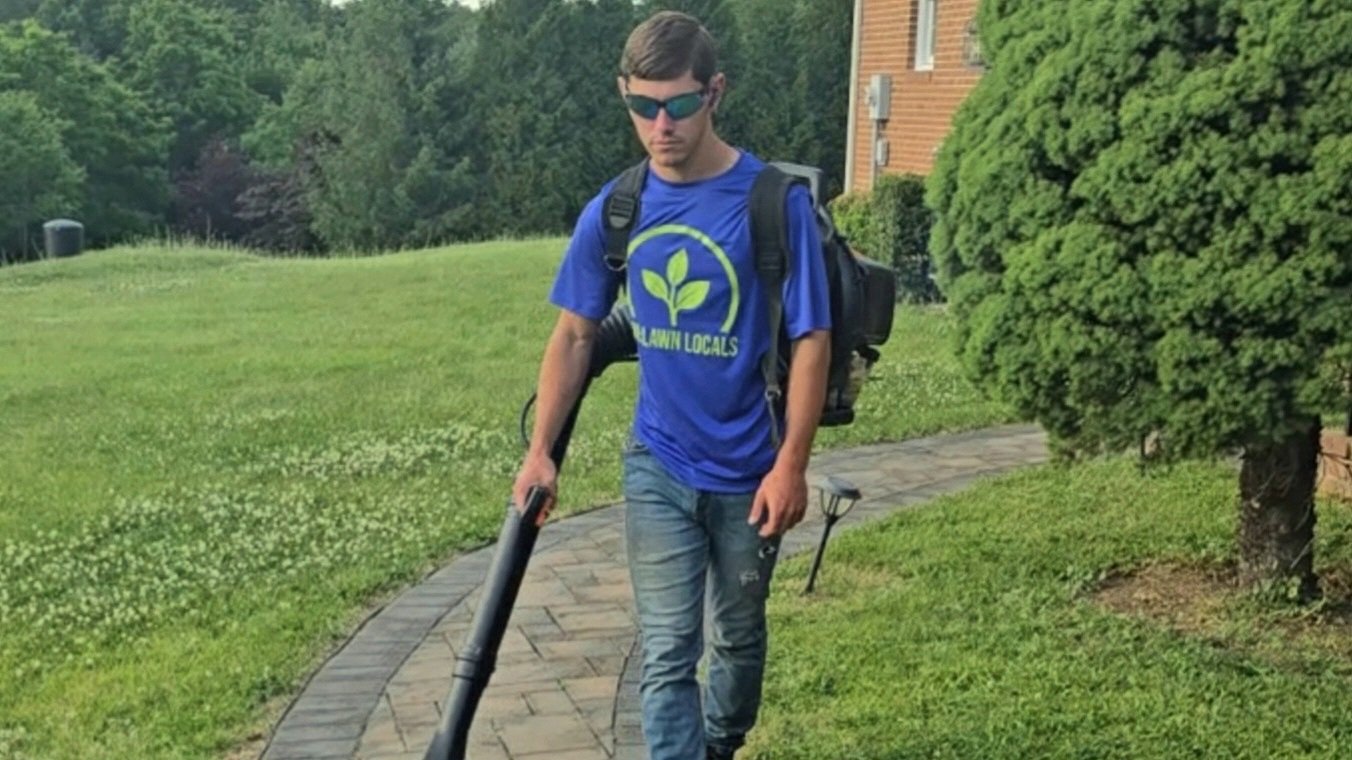 A man wearing sunglasses, a blue Lawn Locals T-shirt, and jeans is using a leaf blower to clean a brick pathway in a green yard with bushes and trees.