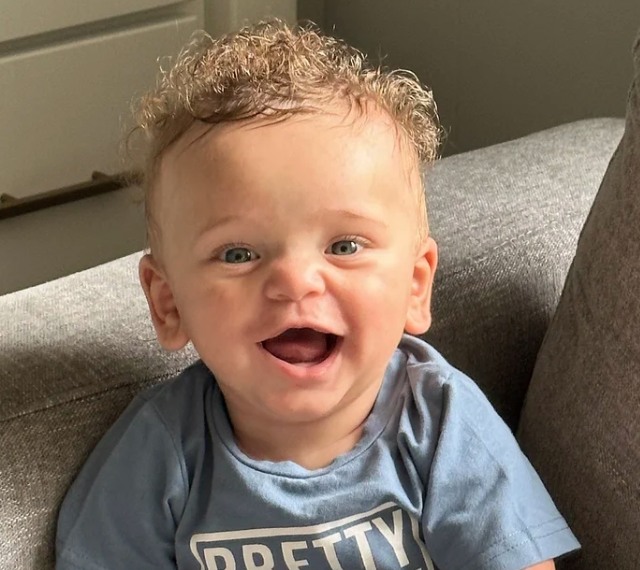 A smiling young boy with curly blonde hair sitting on a gray couch.