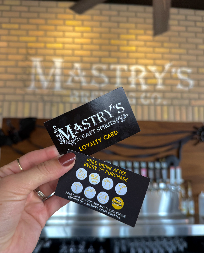Close-up of a hand holding a Mastery's craft spirits loyalty card and a promotional card offering a free drink after every seventh purchase, inside a bar with a brick wall background and the bar's logo projected on the wall.